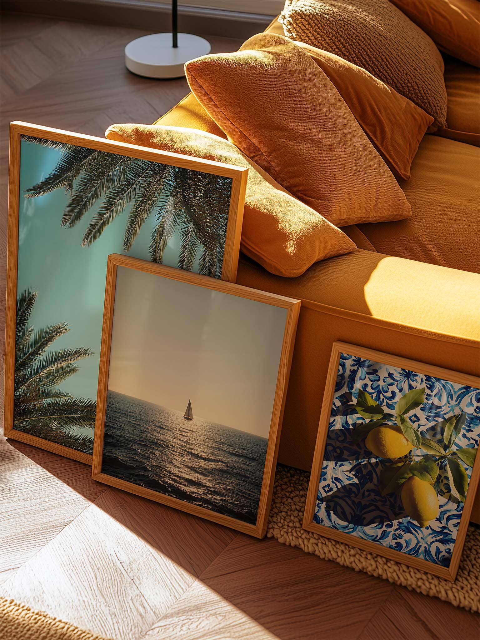 Drei gerahmte Motive mit Palmen vor blauem Himmel, Segelboot auf dem Meer bei Sonnenuntergang und Zitronen auf mediterranen Fliesen, arrangiert neben einem Sofa im warmen Sonnenlicht.