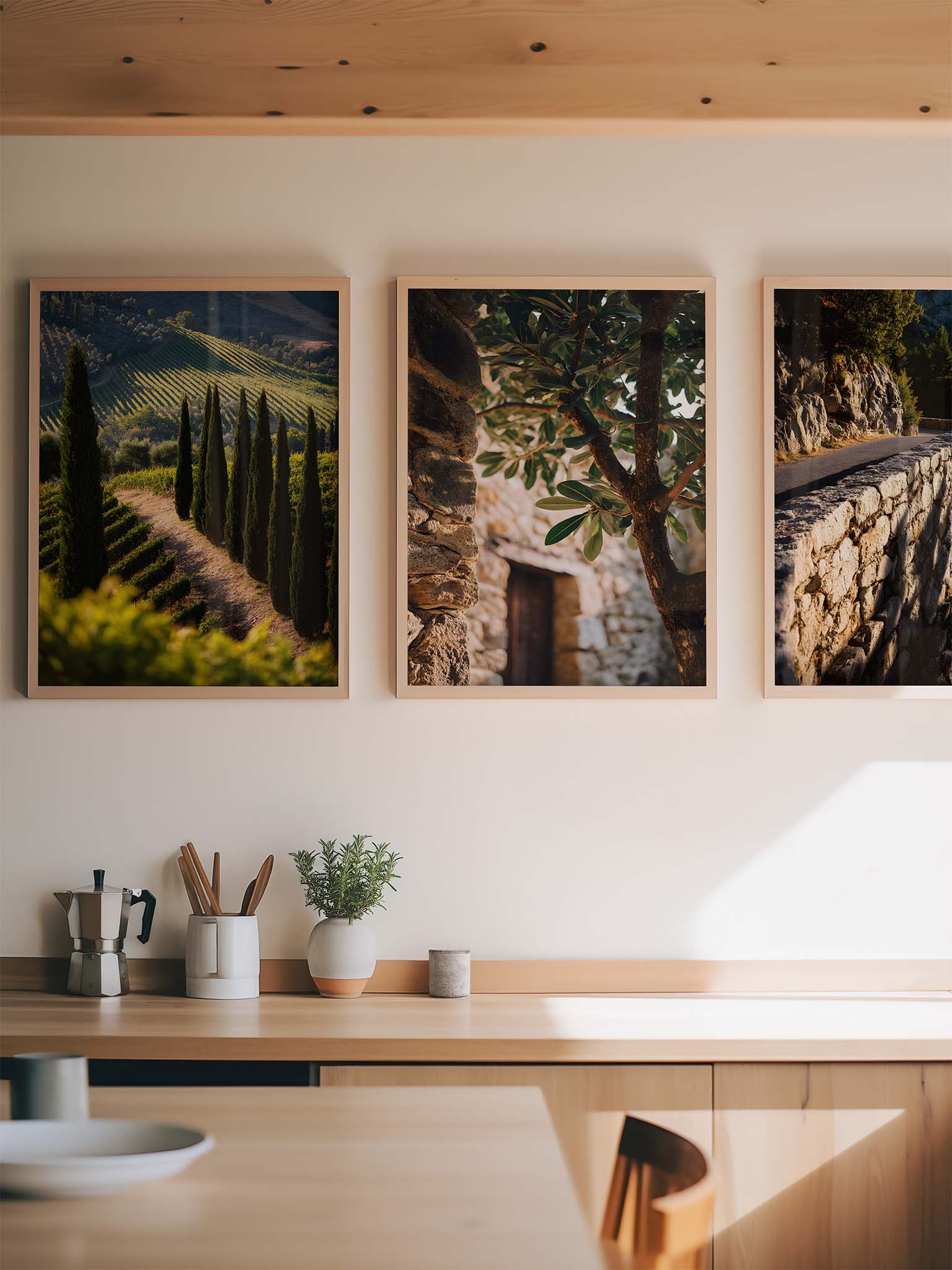 Drei gerahmte Fotografien einer mediterranen Landschaft mit Zypressenallee, Steinmauer und Olivenbaum, arrangiert über einer Holzkommode in einer hellen, minimalistischen Küche.