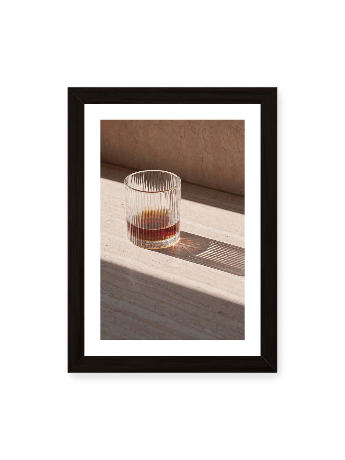 21x30 cm, weißer Galerie-Rand, schwarzer Rahmen – Geripptes Glas mit bernsteinfarbener Flüssigkeit auf heller Steinfläche im direkten Licht.