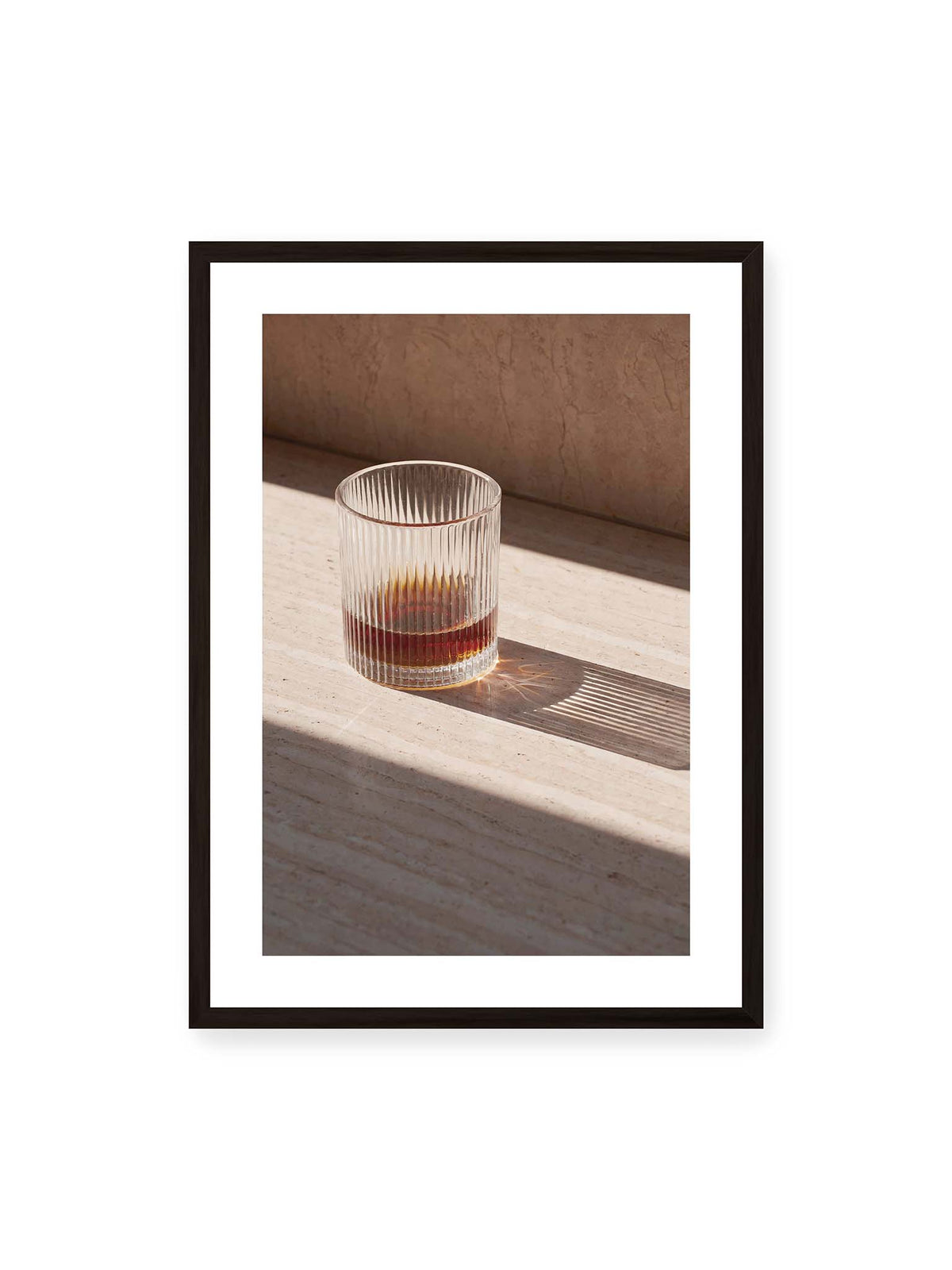 50x70 cm, weißer Galerie-Rand, schwarzer Rahmen – Geripptes Glas mit bernsteinfarbener Flüssigkeit auf heller Steinfläche im direkten Licht.