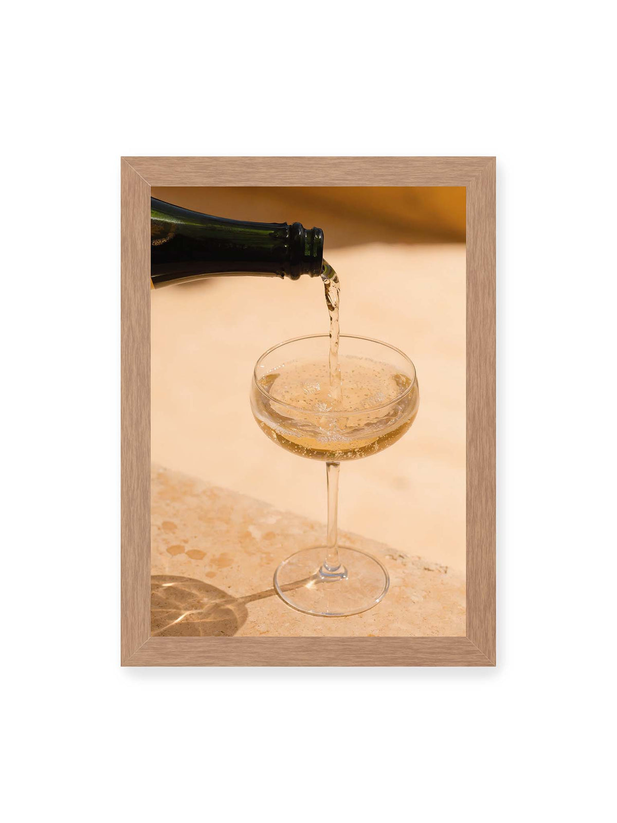 21x30 cm, Eichenrahmen – Perlender Champagner wird aus einer dunklen Flasche in ein Coupéglas auf heller Steinfläche gegossen.