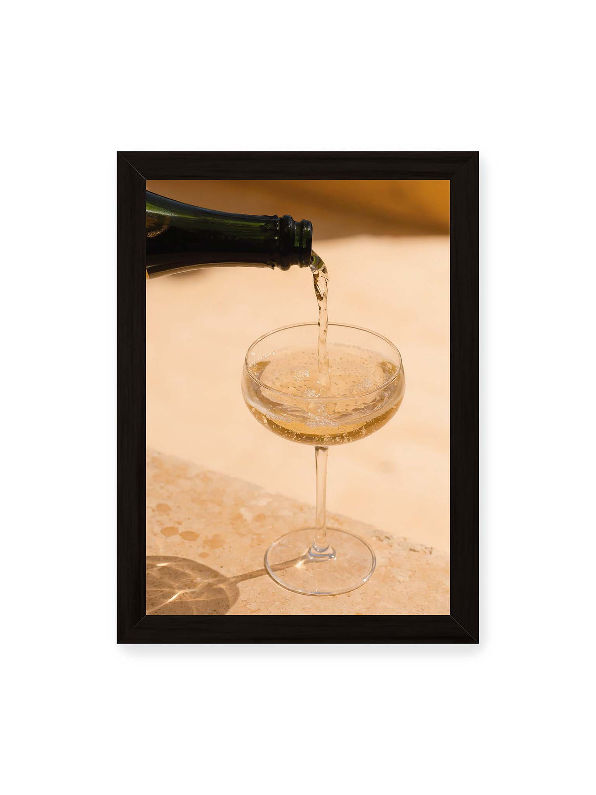 21x30 cm, schwarzer Rahmen – Perlender Champagner wird aus einer dunklen Flasche in ein Coupéglas auf heller Steinfläche gegossen.