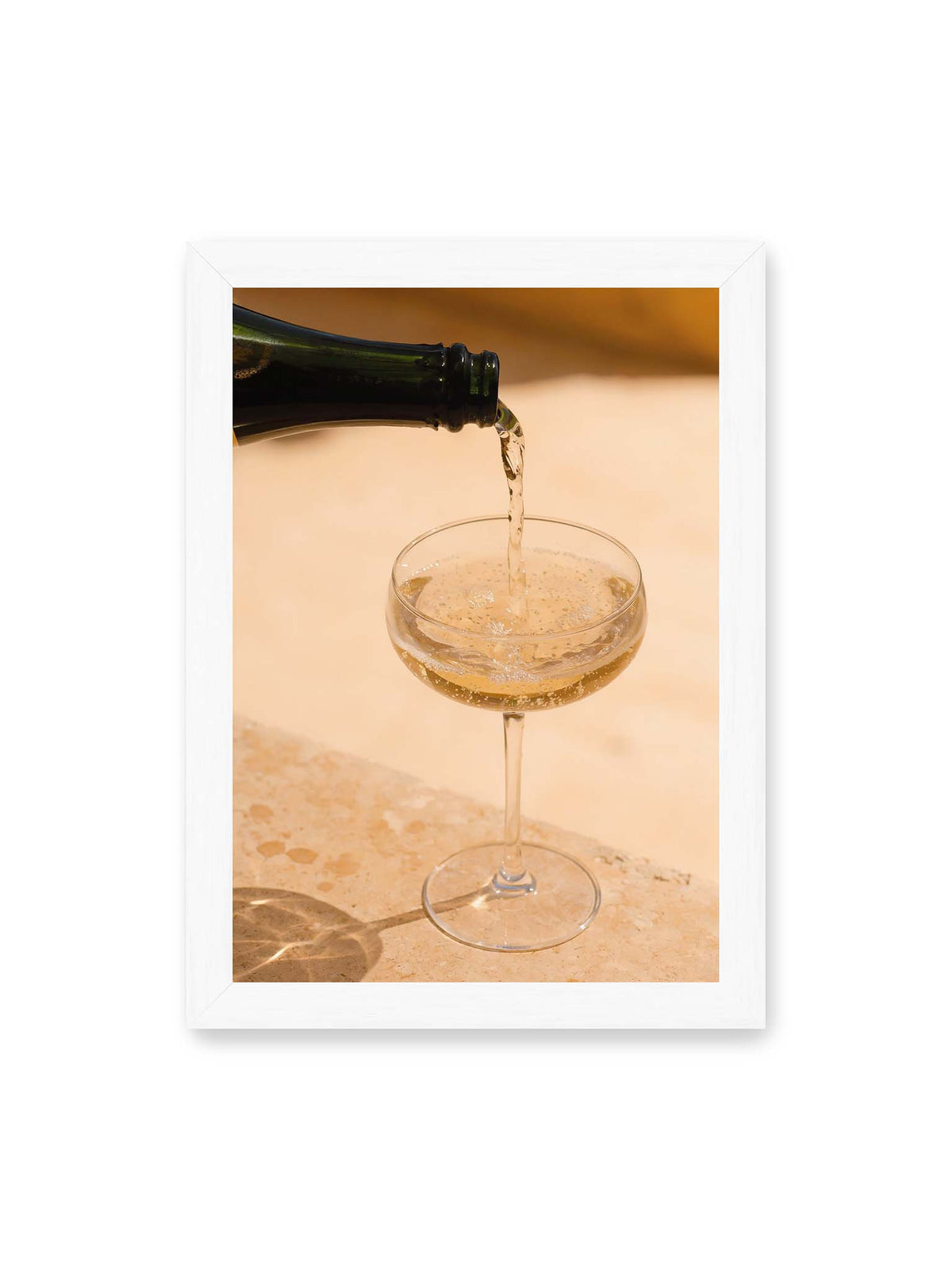 21x30 cm, weißer Rahmen – Perlender Champagner wird aus einer dunklen Flasche in ein Coupéglas auf heller Steinfläche gegossen.
