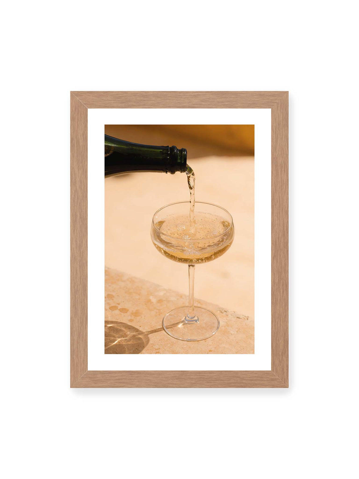 21x30 cm, weißer Galerie-Rand, Eichenrahmen – Perlender Champagner wird aus einer dunklen Flasche in ein Coupéglas auf heller Steinfläche gegossen.