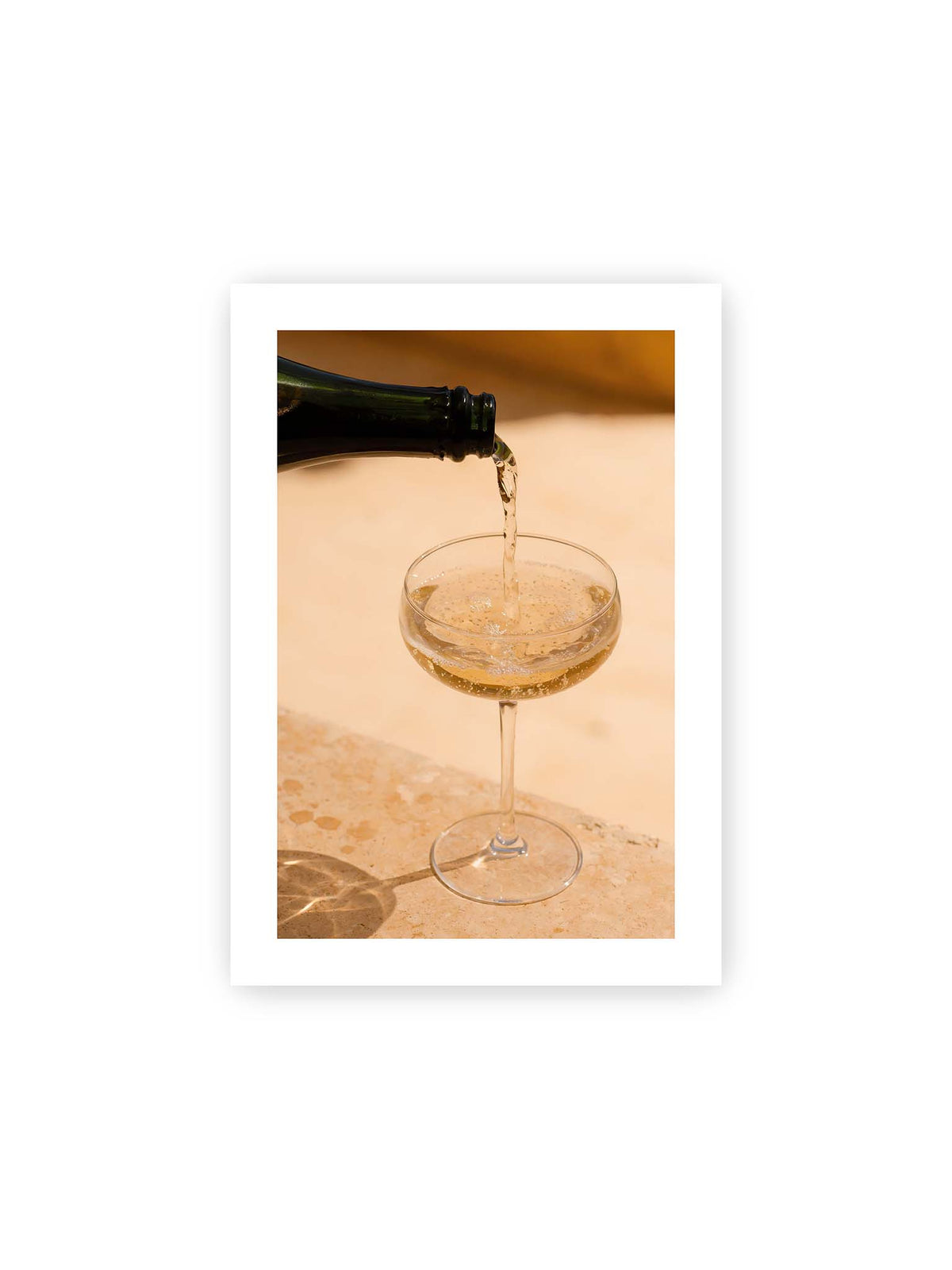 21x30 cm, weißer Galerie-Rand – Perlender Champagner wird aus einer dunklen Flasche in ein Coupéglas auf heller Steinfläche gegossen.