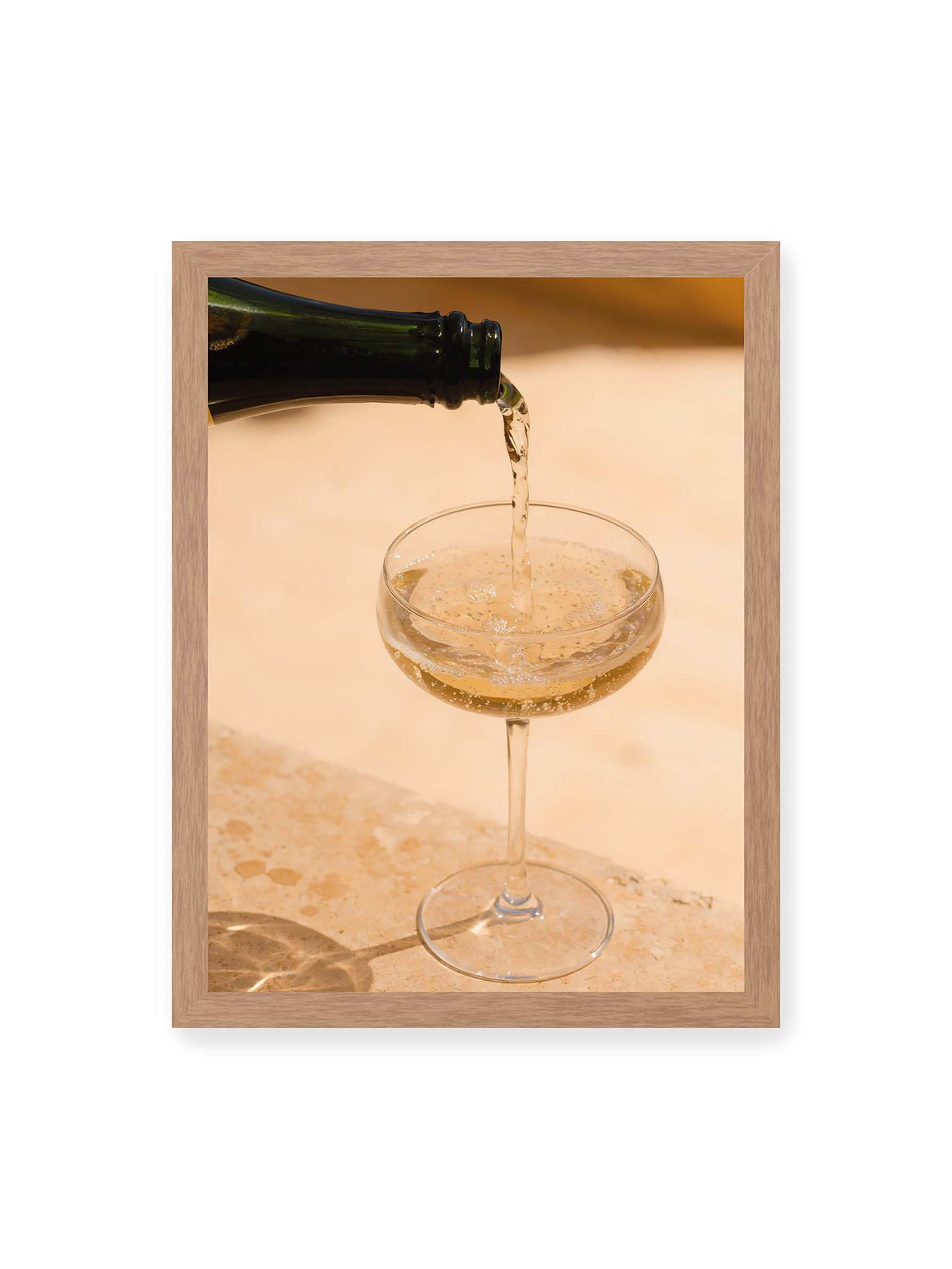 30x40 cm, Eichenrahmen – Perlender Champagner wird aus einer dunklen Flasche in ein Coupéglas auf heller Steinfläche gegossen.