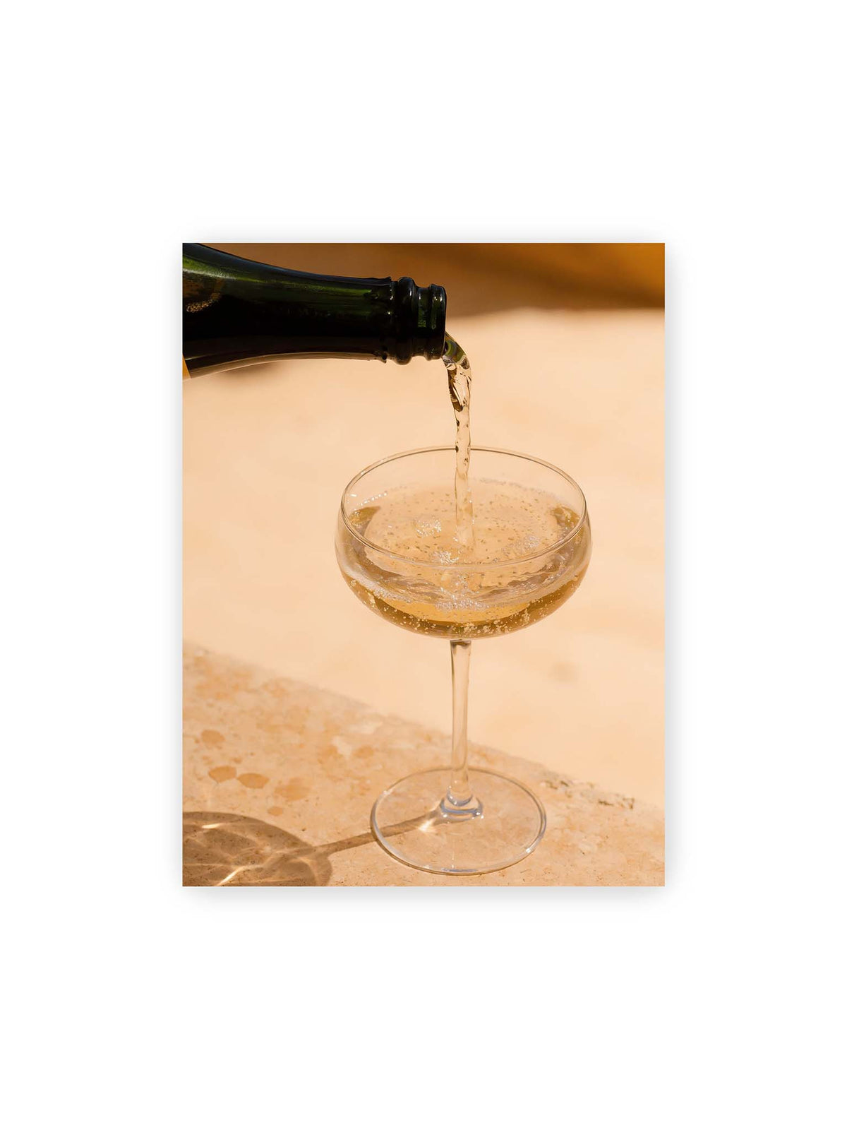 30x40 cm – Perlender Champagner wird aus einer dunklen Flasche in ein Coupéglas auf heller Steinfläche gegossen.