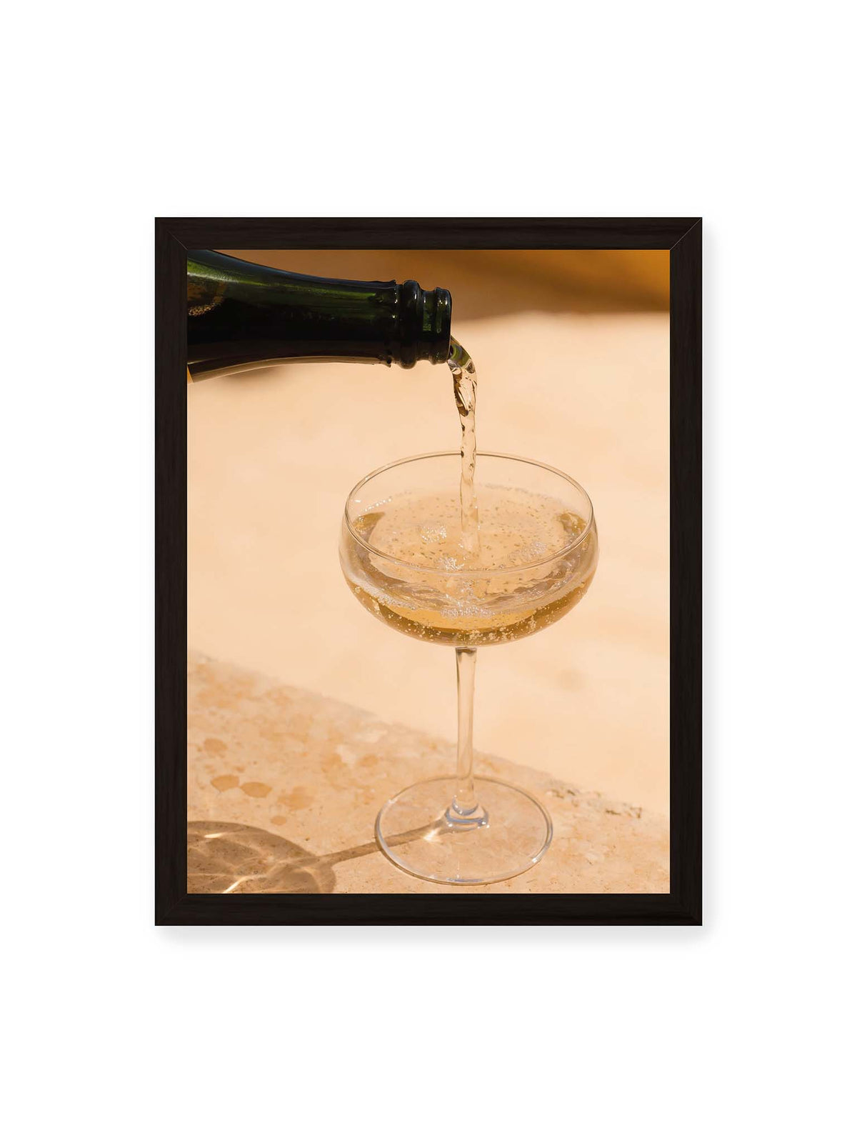 30x40 cm, schwarzer Rahmen – Perlender Champagner wird aus einer dunklen Flasche in ein Coupéglas auf heller Steinfläche gegossen.