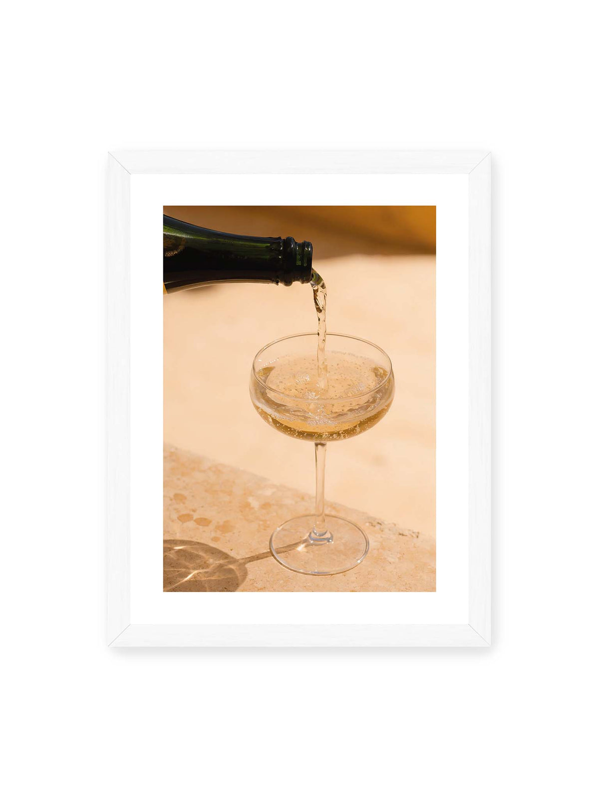 30x40 cm, weißer Galerie-Rand, weißer Rahmen – Perlender Champagner wird aus einer dunklen Flasche in ein Coupéglas auf heller Steinfläche gegossen.