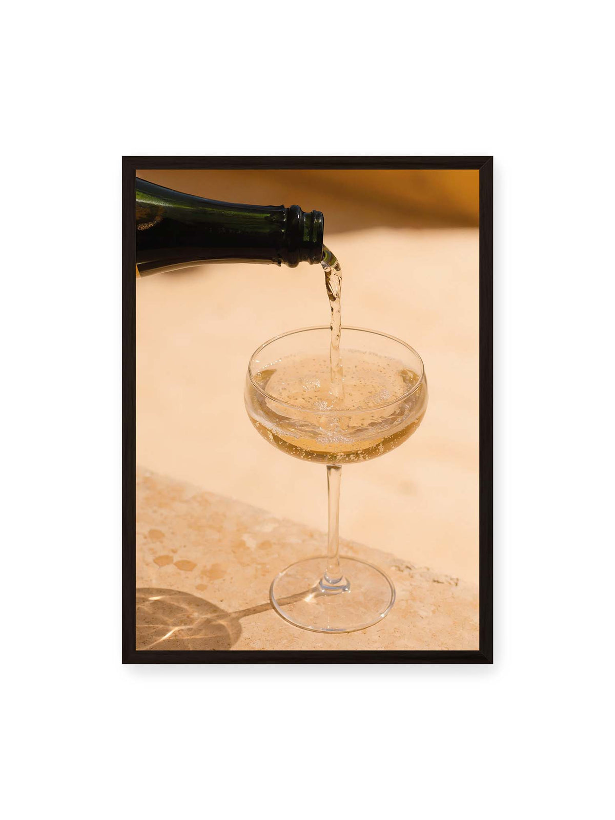 50x70 cm, schwarzer Rahmen – Perlender Champagner wird aus einer dunklen Flasche in ein Coupéglas auf heller Steinfläche gegossen.