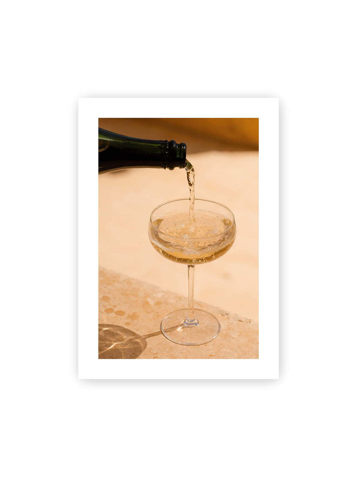 50x70 cm, weißer Galerie-Rand – Perlender Champagner wird aus einer dunklen Flasche in ein Coupéglas auf heller Steinfläche gegossen.