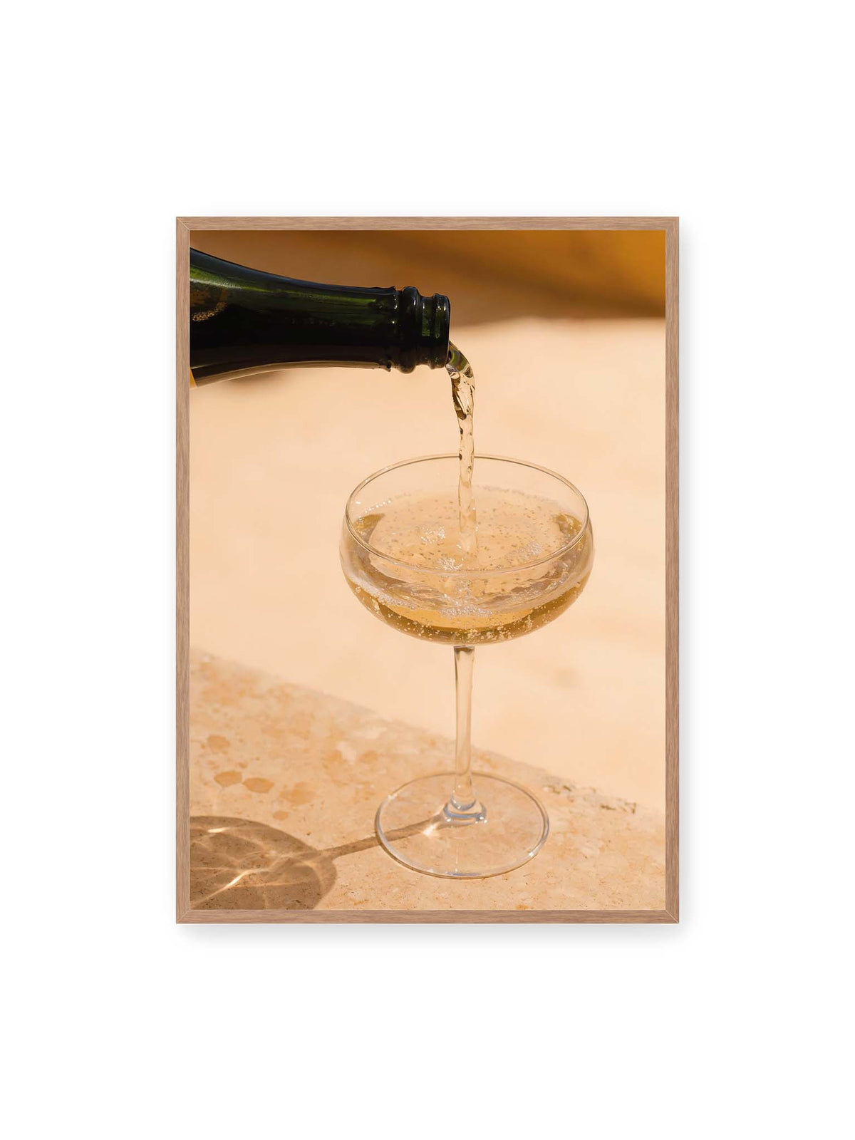 70x100 cm, Eichenrahmen – Perlender Champagner wird aus einer dunklen Flasche in ein Coupéglas auf heller Steinfläche gegossen.