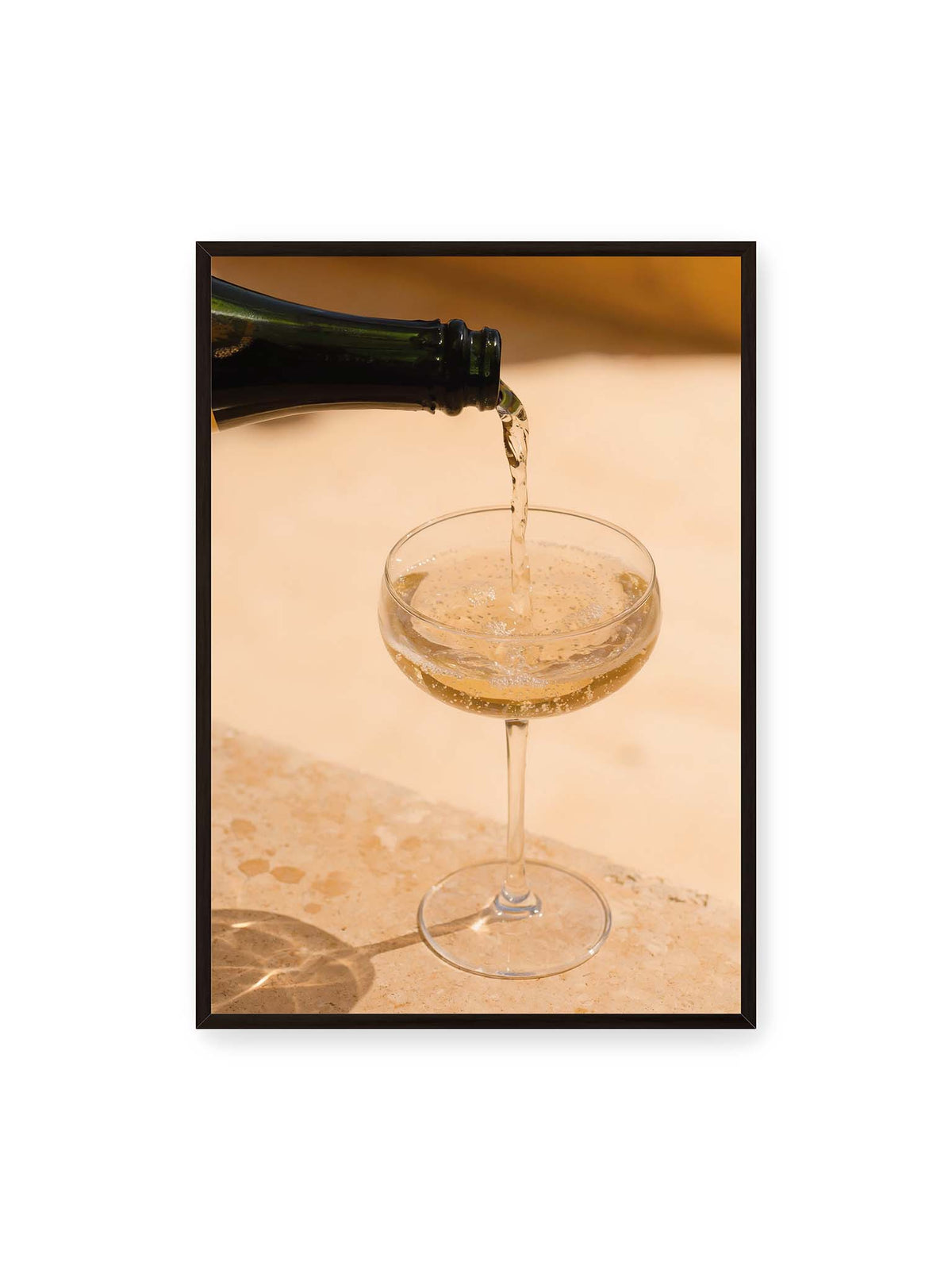 70x100 cm, schwarzer Rahmen – Perlender Champagner wird aus einer dunklen Flasche in ein Coupéglas auf heller Steinfläche gegossen.