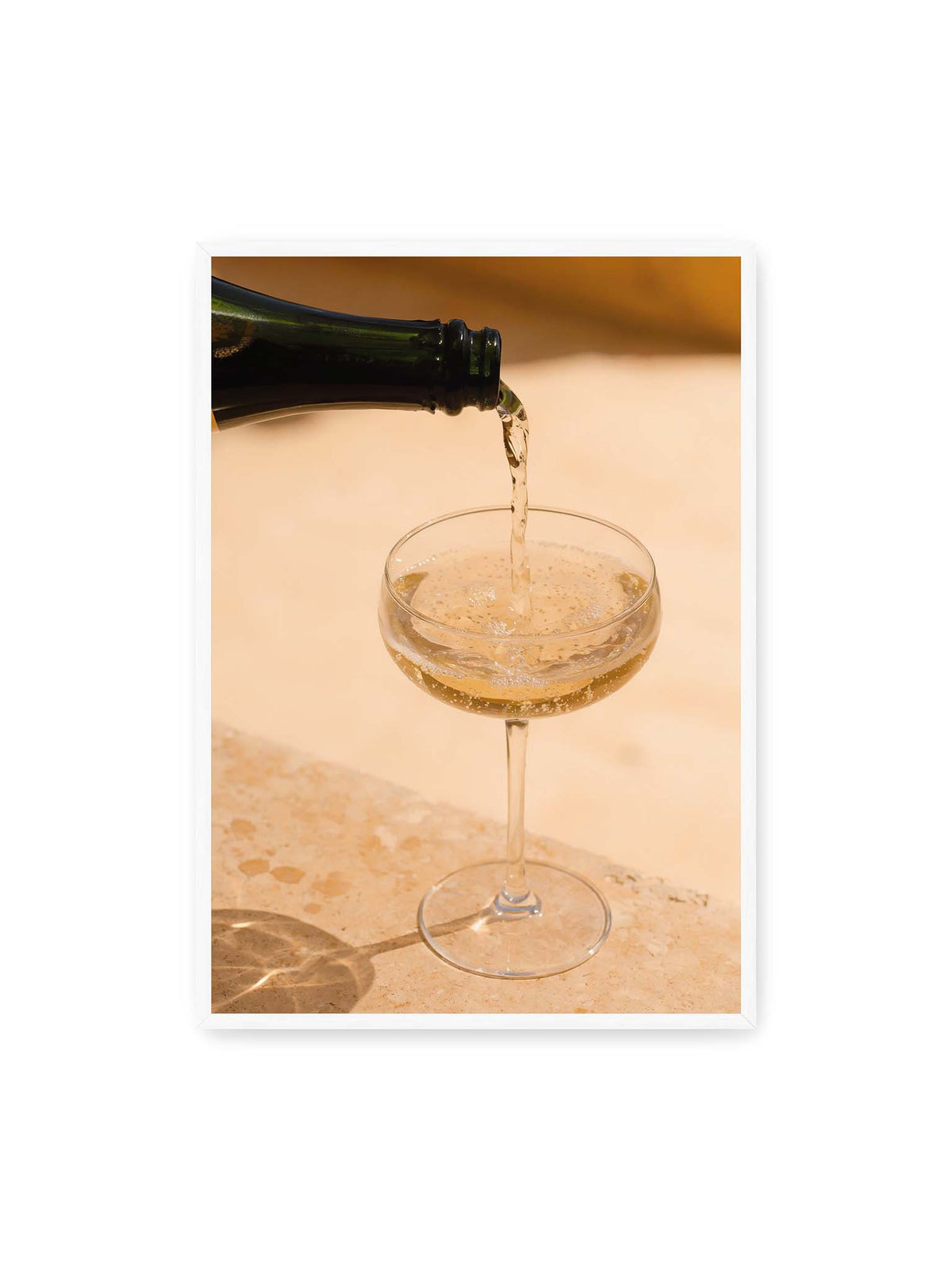 70x100 cm, weißer Rahmen – Perlender Champagner wird aus einer dunklen Flasche in ein Coupéglas auf heller Steinfläche gegossen.
