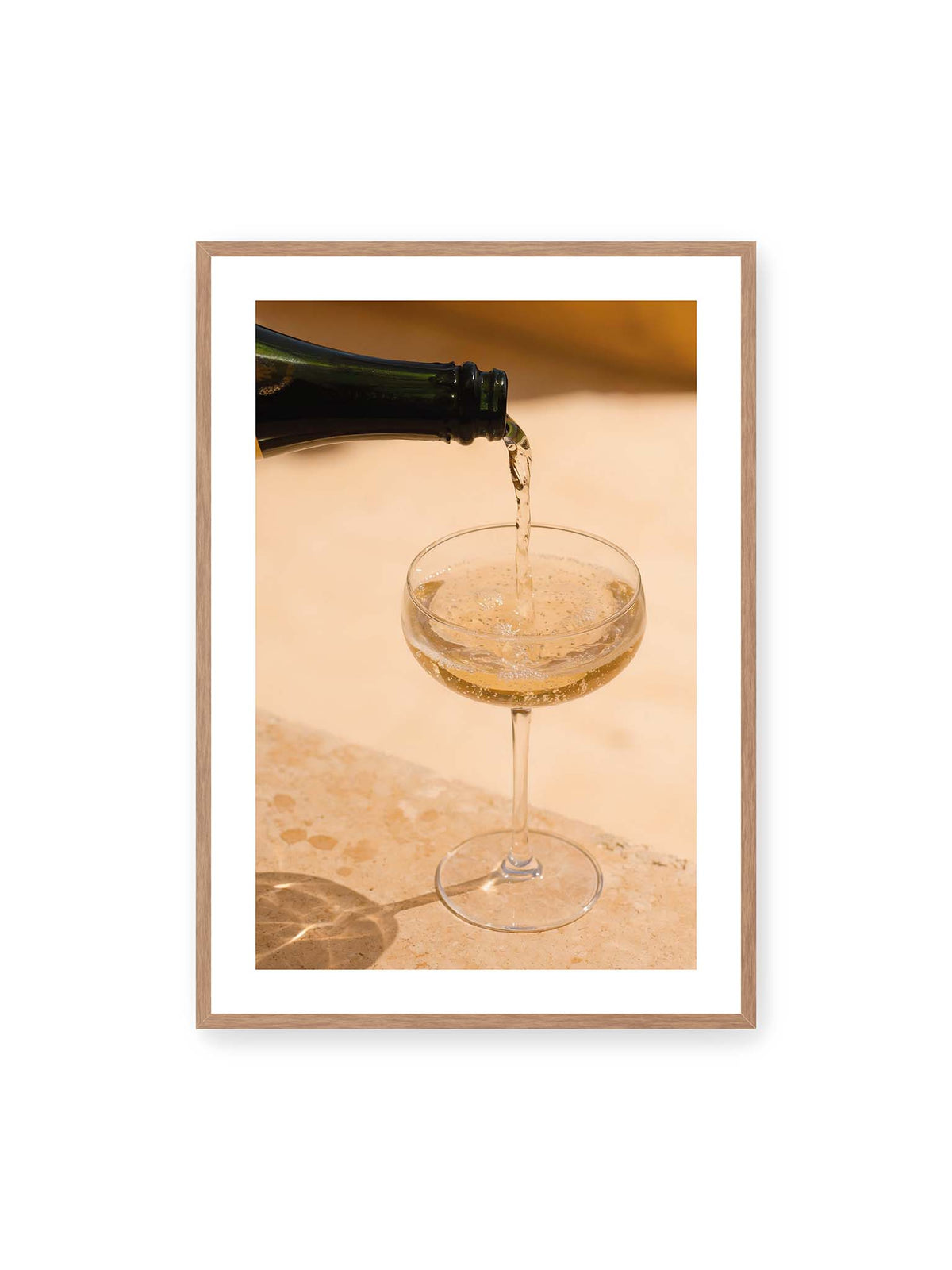 70x100 cm, weißer Galerie-Rand, Eichenrahmen – Perlender Champagner wird aus einer dunklen Flasche in ein Coupéglas auf heller Steinfläche gegossen.