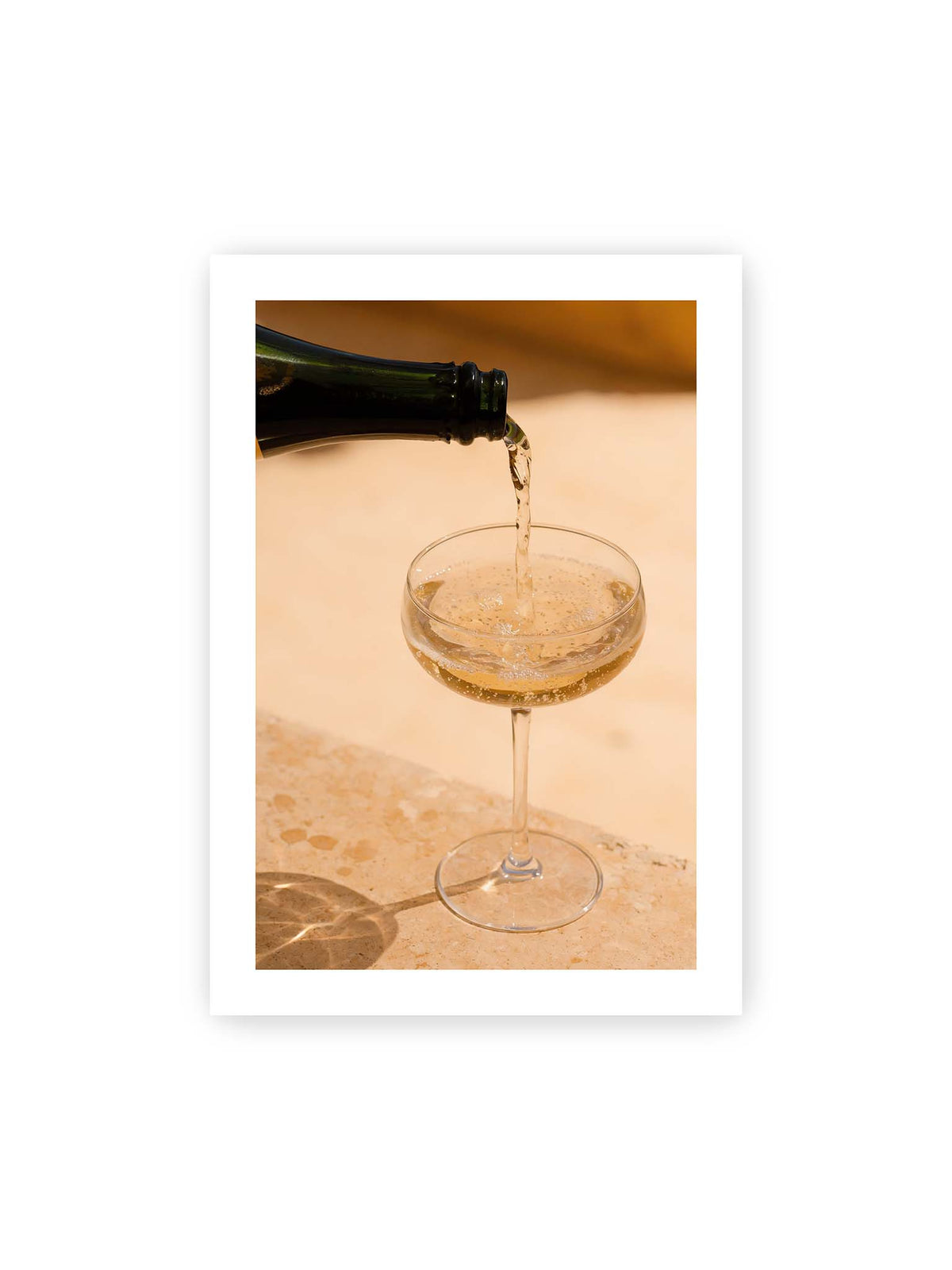 70x100 cm, weißer Galerie-Rand – Perlender Champagner wird aus einer dunklen Flasche in ein Coupéglas auf heller Steinfläche gegossen.
