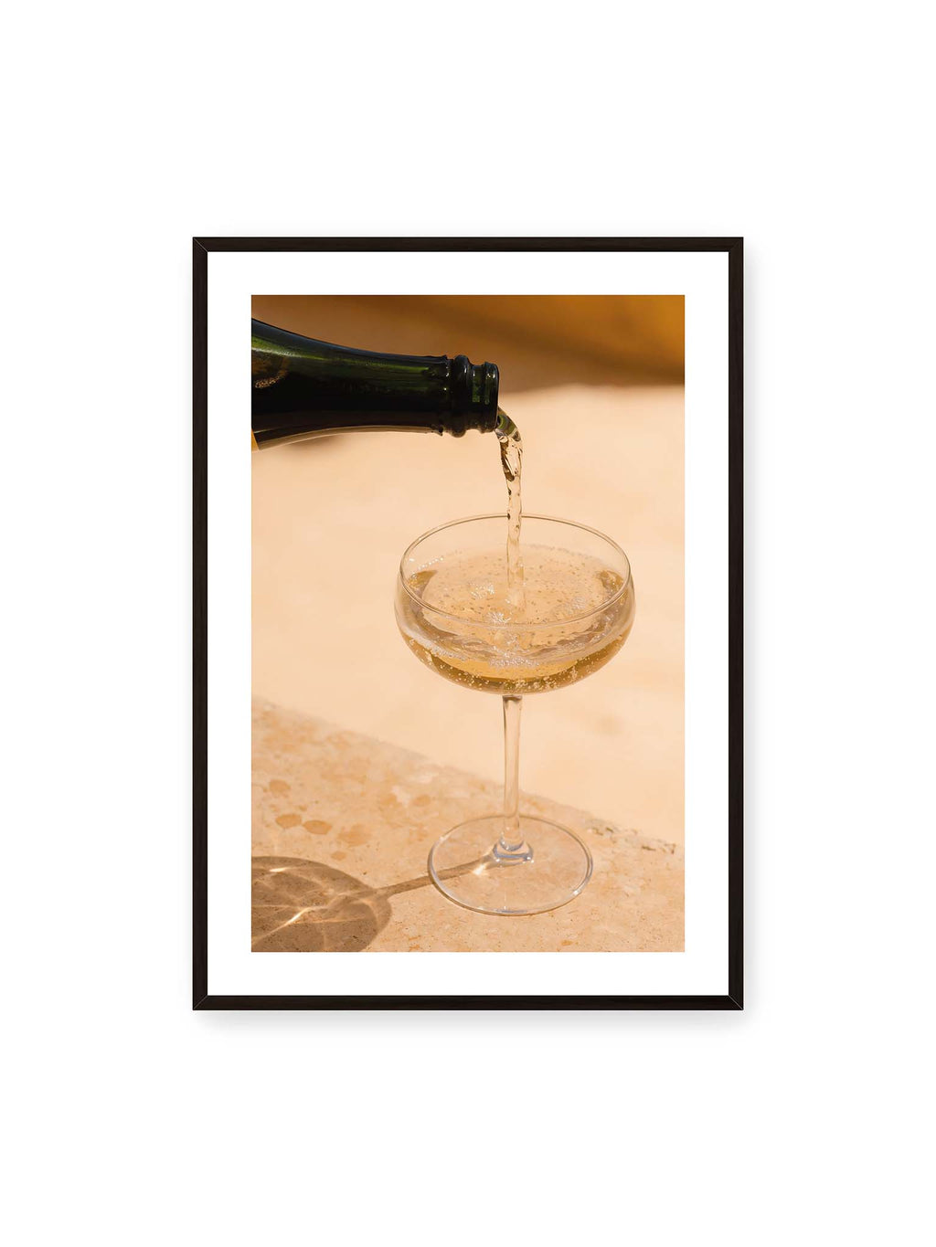 70x100 cm, weißer Galerie-Rand, schwarzer Rahmen – Perlender Champagner wird aus einer dunklen Flasche in ein Coupéglas auf heller Steinfläche gegossen.