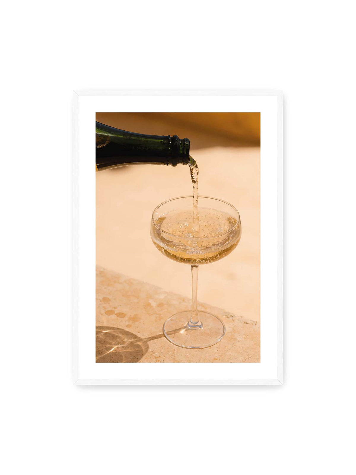 70x100 cm, weißer Galerie-Rand, weißer Rahmen – Perlender Champagner wird aus einer dunklen Flasche in ein Coupéglas auf heller Steinfläche gegossen.