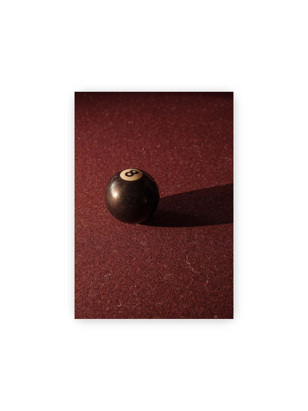 21x30 cm – Schwarze Billardkugel auf tiefroter Spielfläche mit langem Schatten im seitlichen Licht.