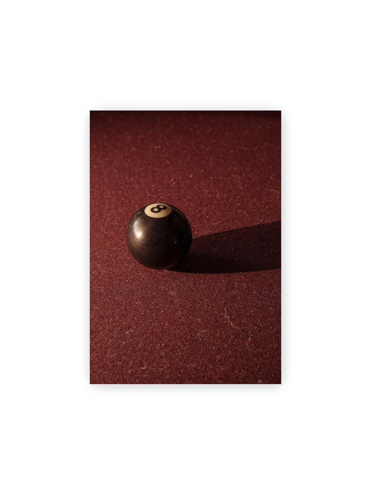 21x30 cm – Schwarze Billardkugel auf tiefroter Spielfläche mit langem Schatten im seitlichen Licht.