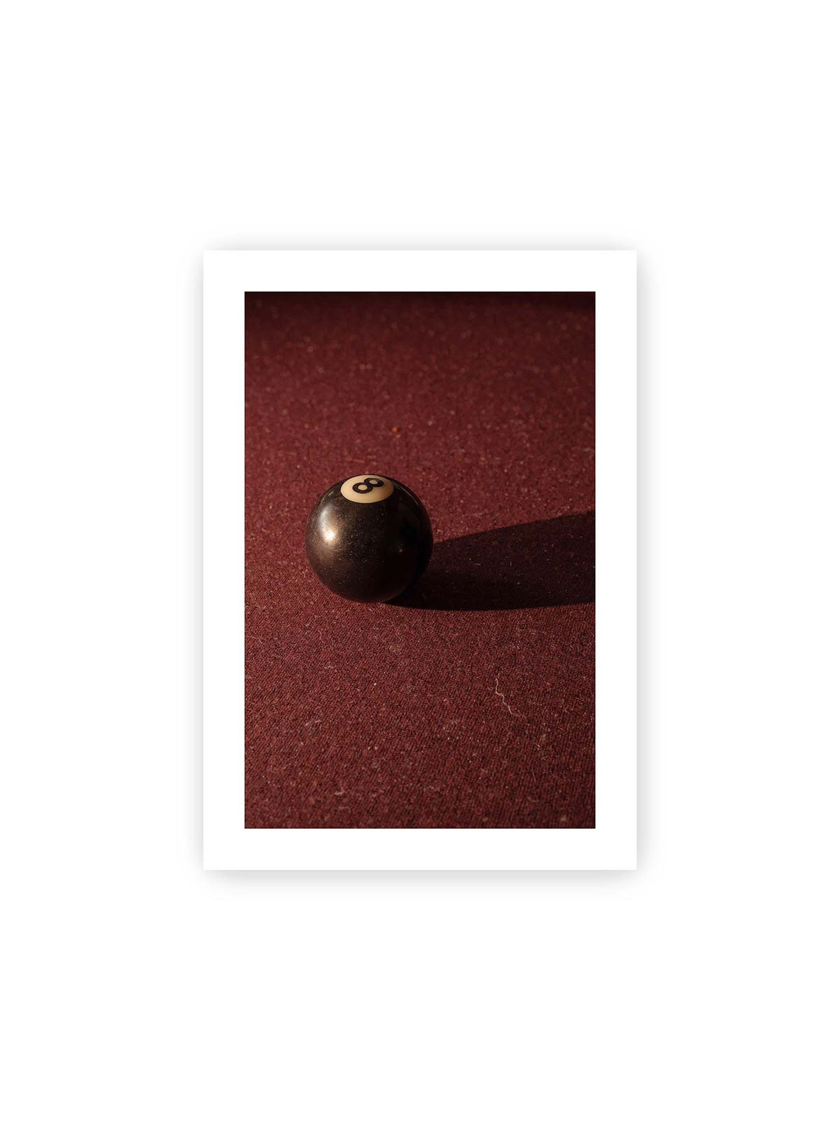 21x30 cm, weißer Galerie-Rand – Schwarze Billardkugel auf tiefroter Spielfläche mit langem Schatten im seitlichen Licht.