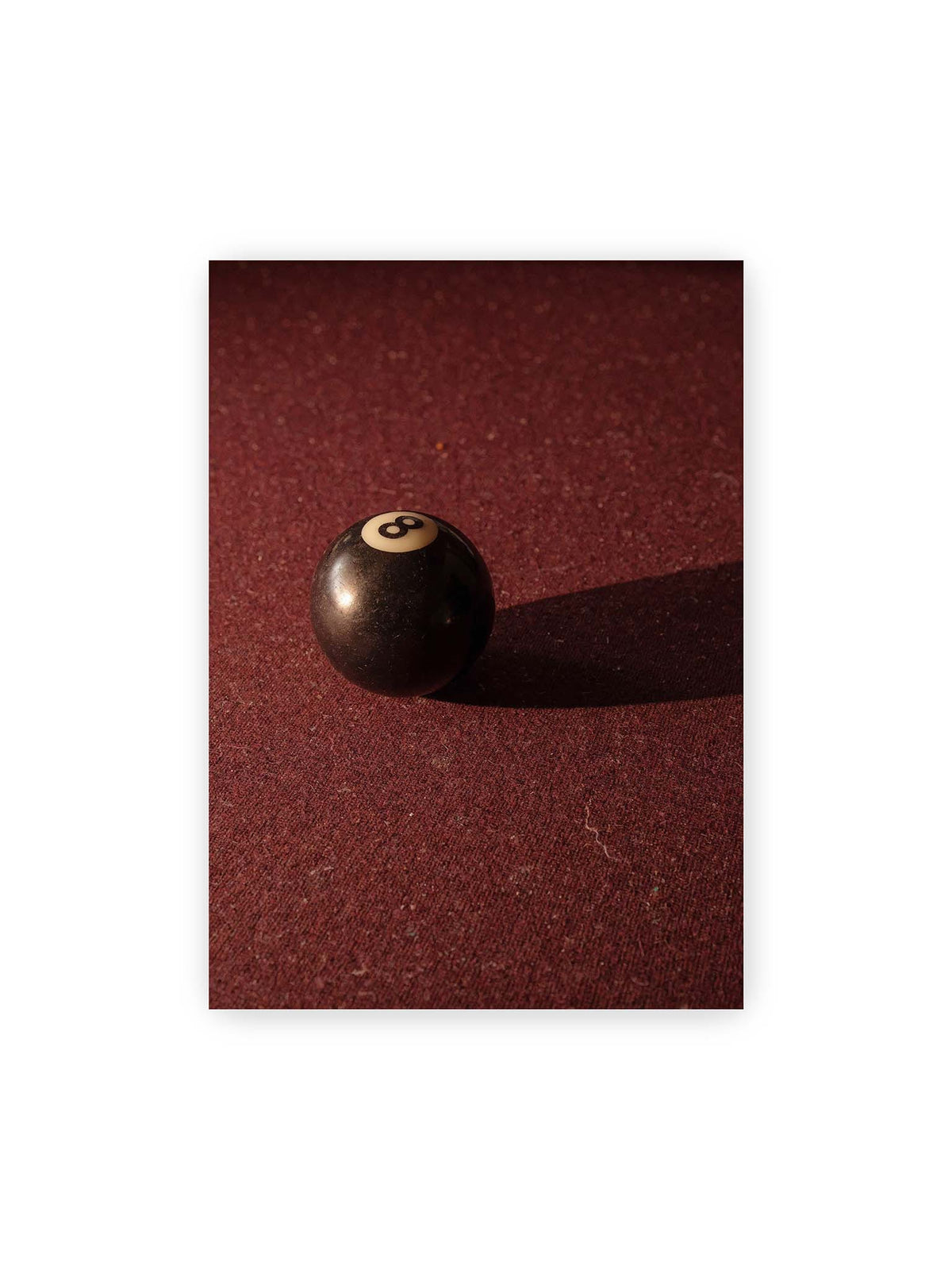 50x70 cm – Schwarze Billardkugel auf tiefroter Spielfläche mit langem Schatten im seitlichen Licht.
