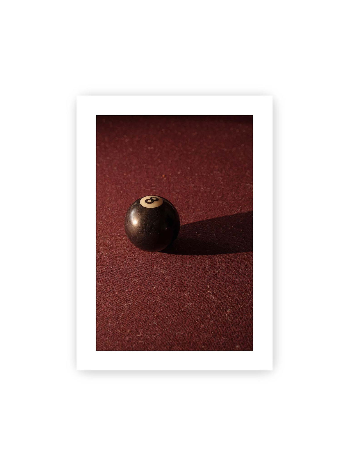50x70 cm, weißer Galerie-Rand – Schwarze Billardkugel auf tiefroter Spielfläche mit langem Schatten im seitlichen Licht.
