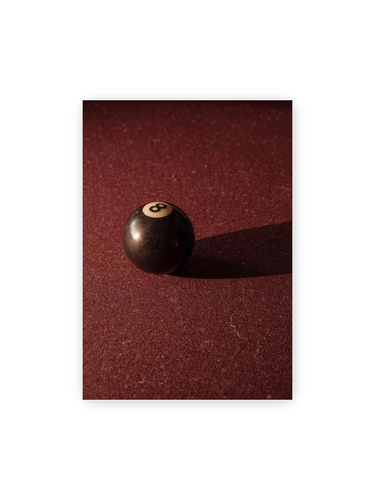 70x100 cm – Schwarze Billardkugel auf tiefroter Spielfläche mit langem Schatten im seitlichen Licht.