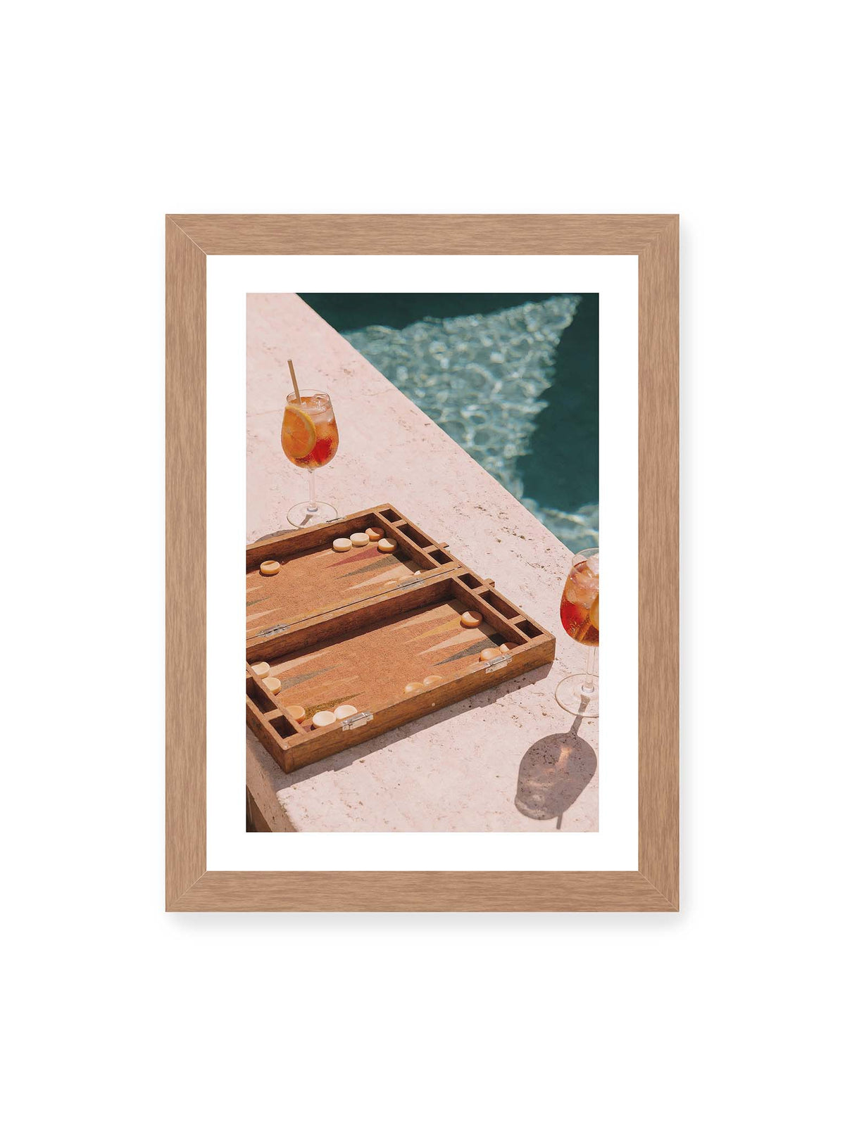 21x30 cm, weißer Galerie-Rand, Eichenrahmen – Geöffnetes Backgammonbrett mit zwei orangefarbenen Drinks auf heller Steinfläche neben blauem Wasser.