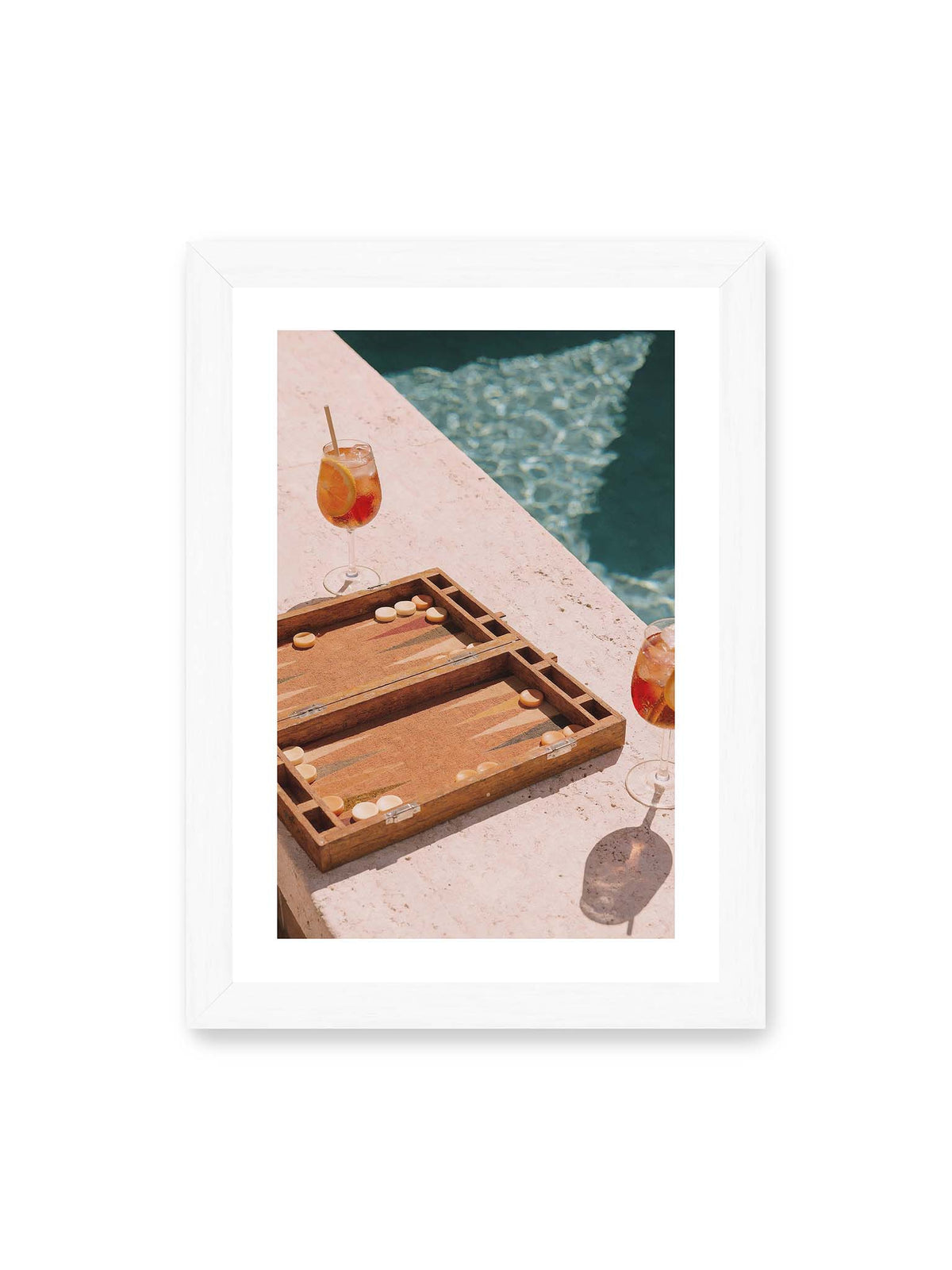 21x30 cm, weißer Galerie-Rand, weißer Rahmen – Geöffnetes Backgammonbrett mit zwei orangefarbenen Drinks auf heller Steinfläche neben blauem Wasser.