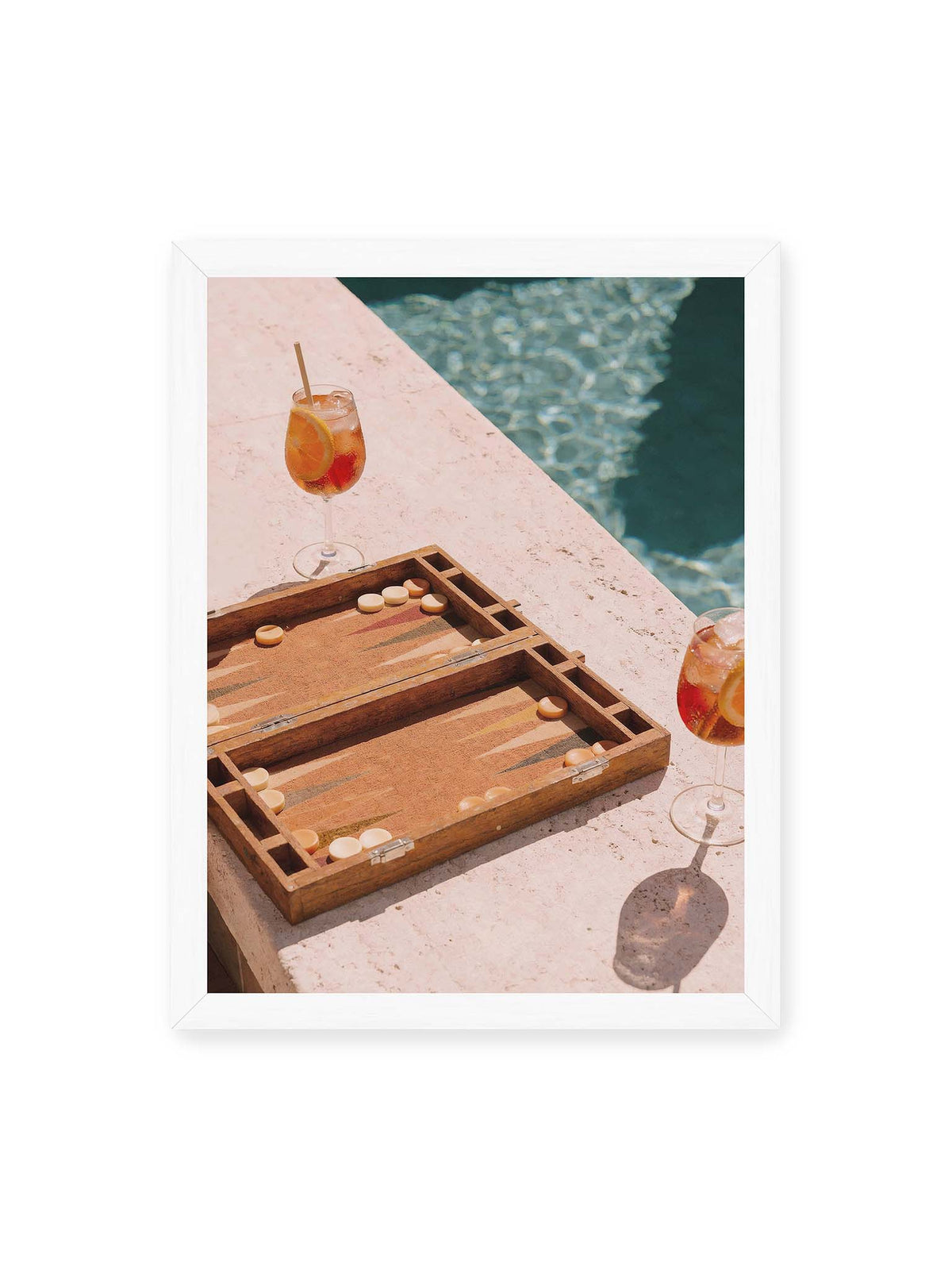 30x40 cm, weißer Rahmen – Geöffnetes Backgammonbrett mit zwei orangefarbenen Drinks auf heller Steinfläche neben blauem Wasser.