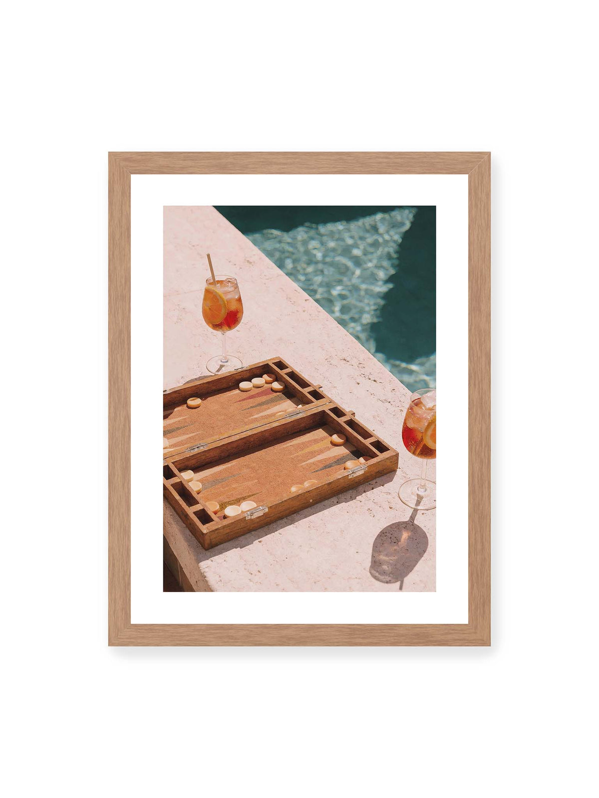 30x40 cm, weißer Galerie-Rand, Eichenrahmen – Geöffnetes Backgammonbrett mit zwei orangefarbenen Drinks auf heller Steinfläche neben blauem Wasser.
