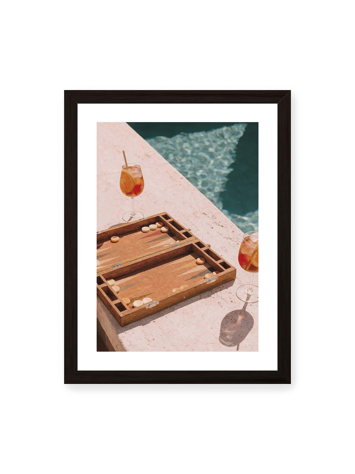 30x40 cm, weißer Galerie-Rand, schwarzer Rahmen – Geöffnetes Backgammonbrett mit zwei orangefarbenen Drinks auf heller Steinfläche neben blauem Wasser.