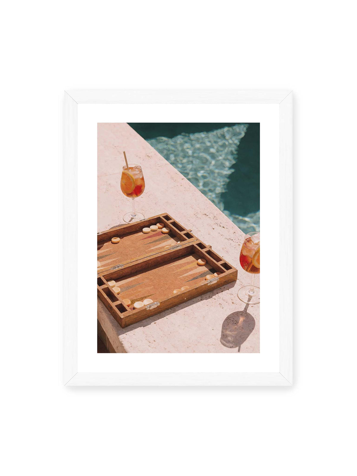30x40 cm, weißer Galerie-Rand, weißer Rahmen – Geöffnetes Backgammonbrett mit zwei orangefarbenen Drinks auf heller Steinfläche neben blauem Wasser.