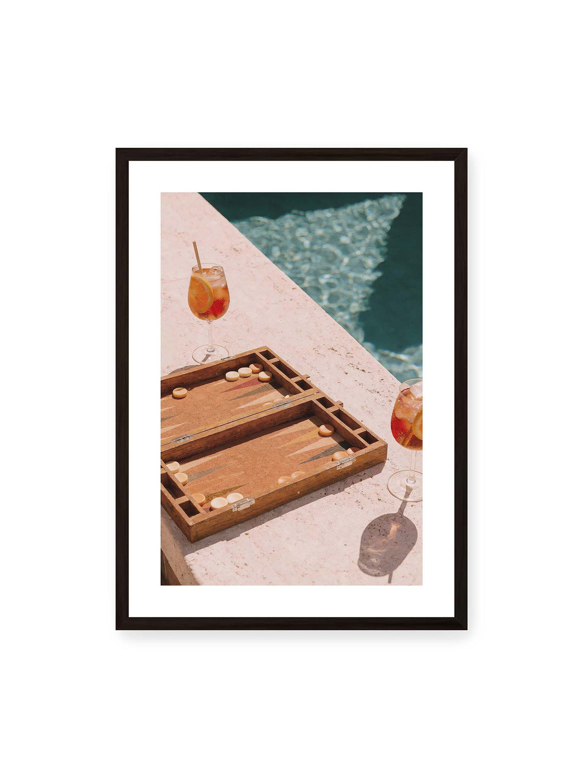 50x70 cm, weißer Galerie-Rand, schwarzer Rahmen – Geöffnetes Backgammonbrett mit zwei orangefarbenen Drinks auf heller Steinfläche neben blauem Wasser.