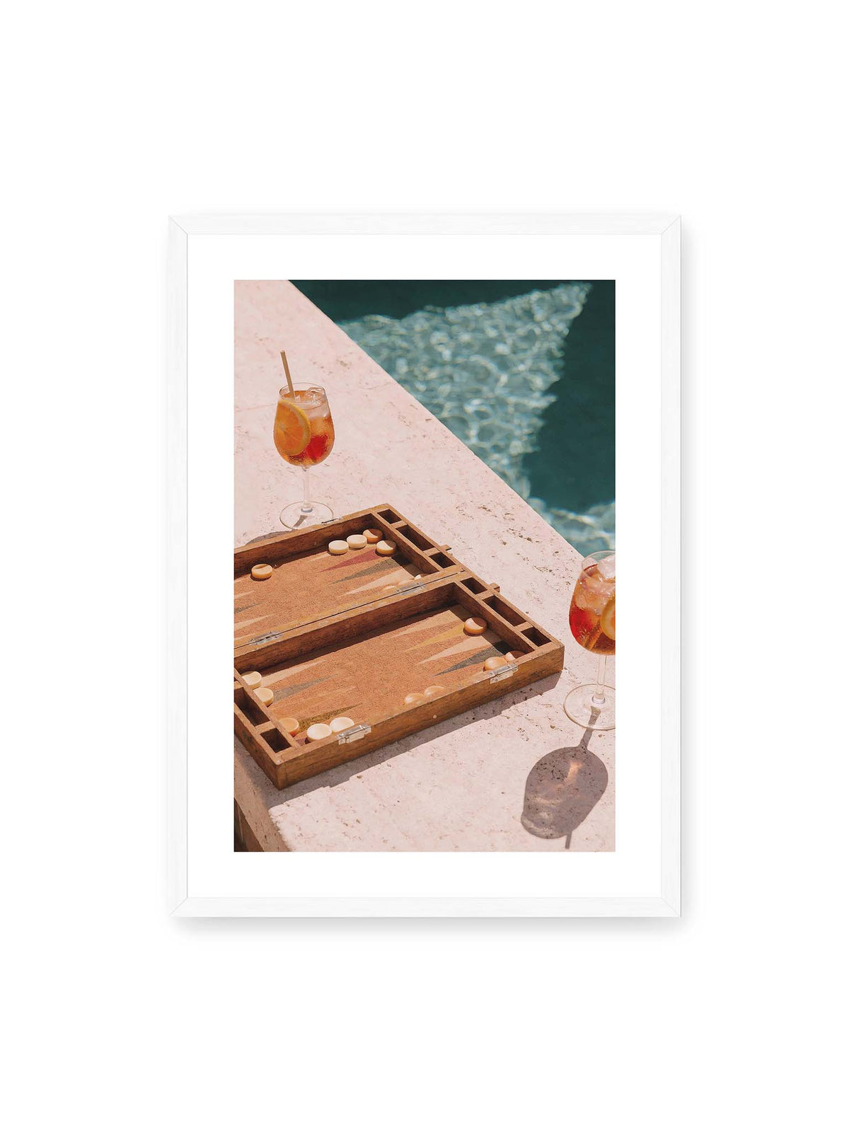50x70 cm, weißer Galerie-Rand, weißer Rahmen – Geöffnetes Backgammonbrett mit zwei orangefarbenen Drinks auf heller Steinfläche neben blauem Wasser.