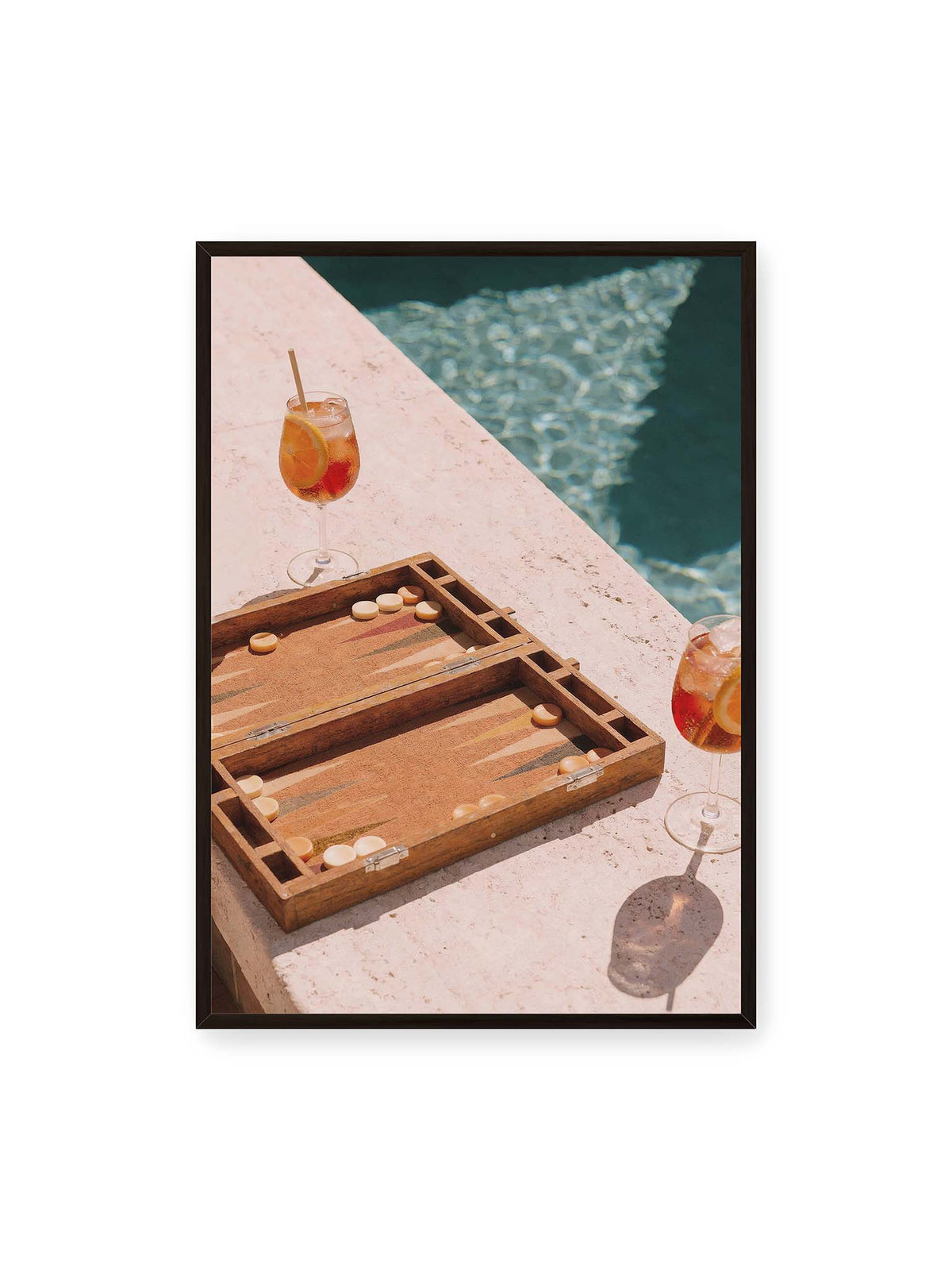 70x100 cm, schwarzer Rahmen – Geöffnetes Backgammonbrett mit zwei orangefarbenen Drinks auf heller Steinfläche neben blauem Wasser.