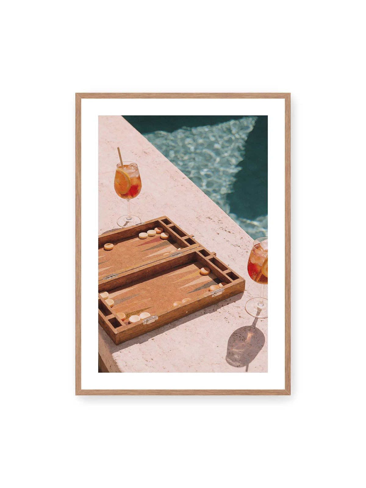 70x100 cm, weißer Galerie-Rand, Eichenrahmen – Geöffnetes Backgammonbrett mit zwei orangefarbenen Drinks auf heller Steinfläche neben blauem Wasser.