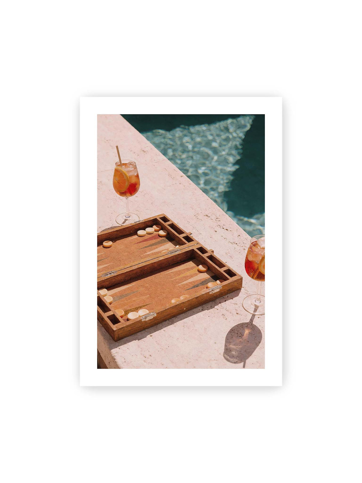 70x100 cm, weißer Galerie-Rand – Geöffnetes Backgammonbrett mit zwei orangefarbenen Drinks auf heller Steinfläche neben blauem Wasser.