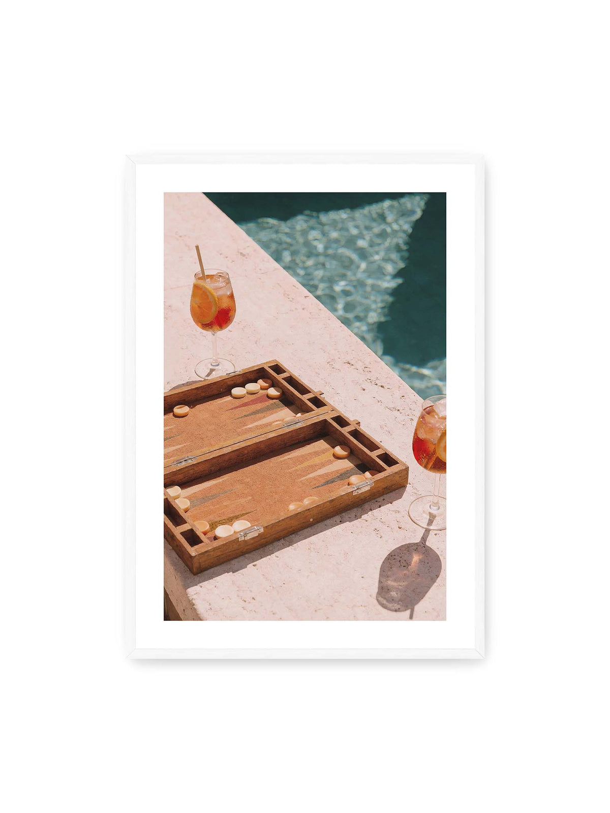 70x100 cm, weißer Galerie-Rand, weißer Rahmen – Geöffnetes Backgammonbrett mit zwei orangefarbenen Drinks auf heller Steinfläche neben blauem Wasser.