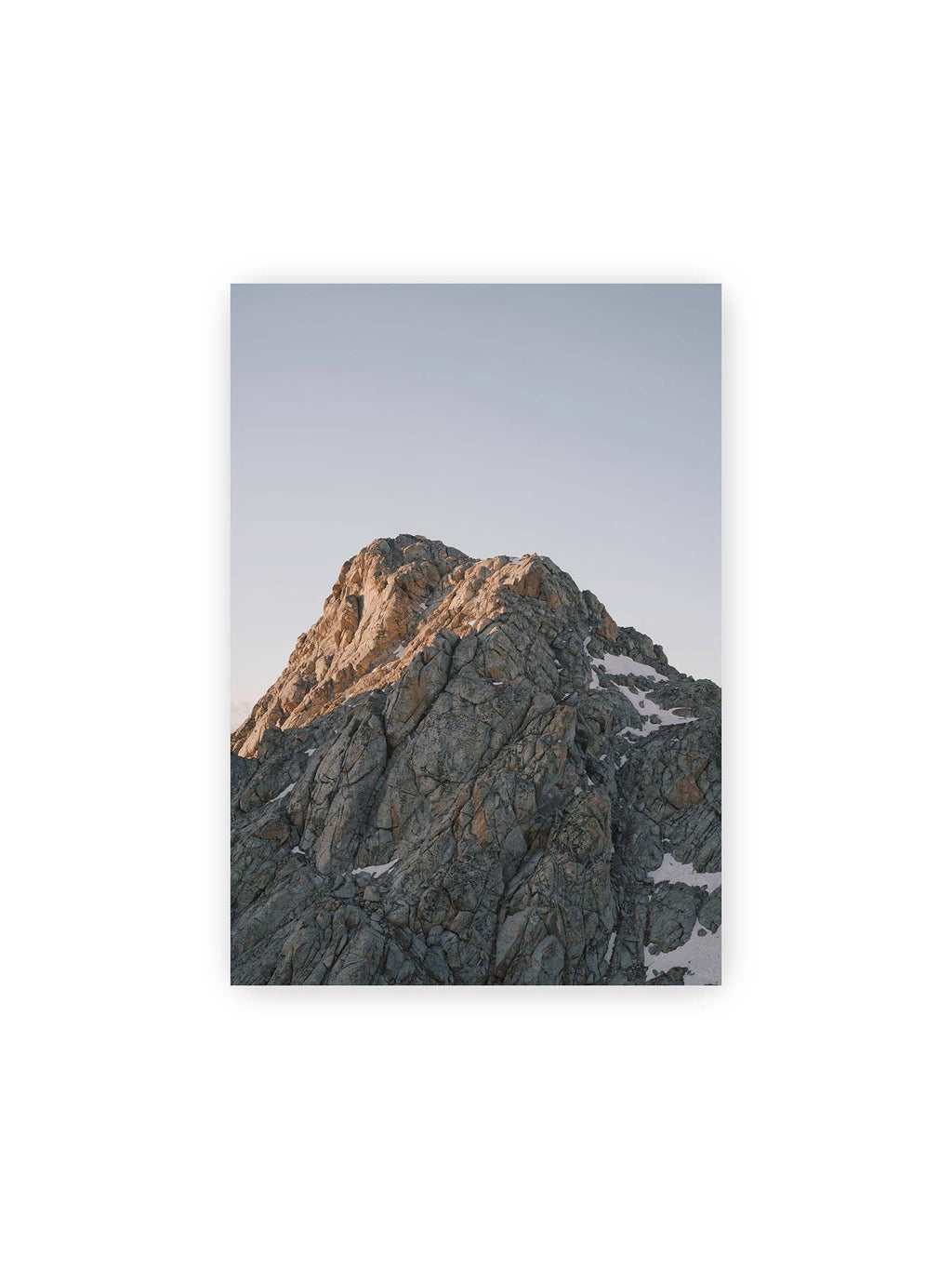 21x30 cm – Massiver grauer Felsgipfel mit kleinen Schneefeldern vor einem weiten blassblauen Himmel.