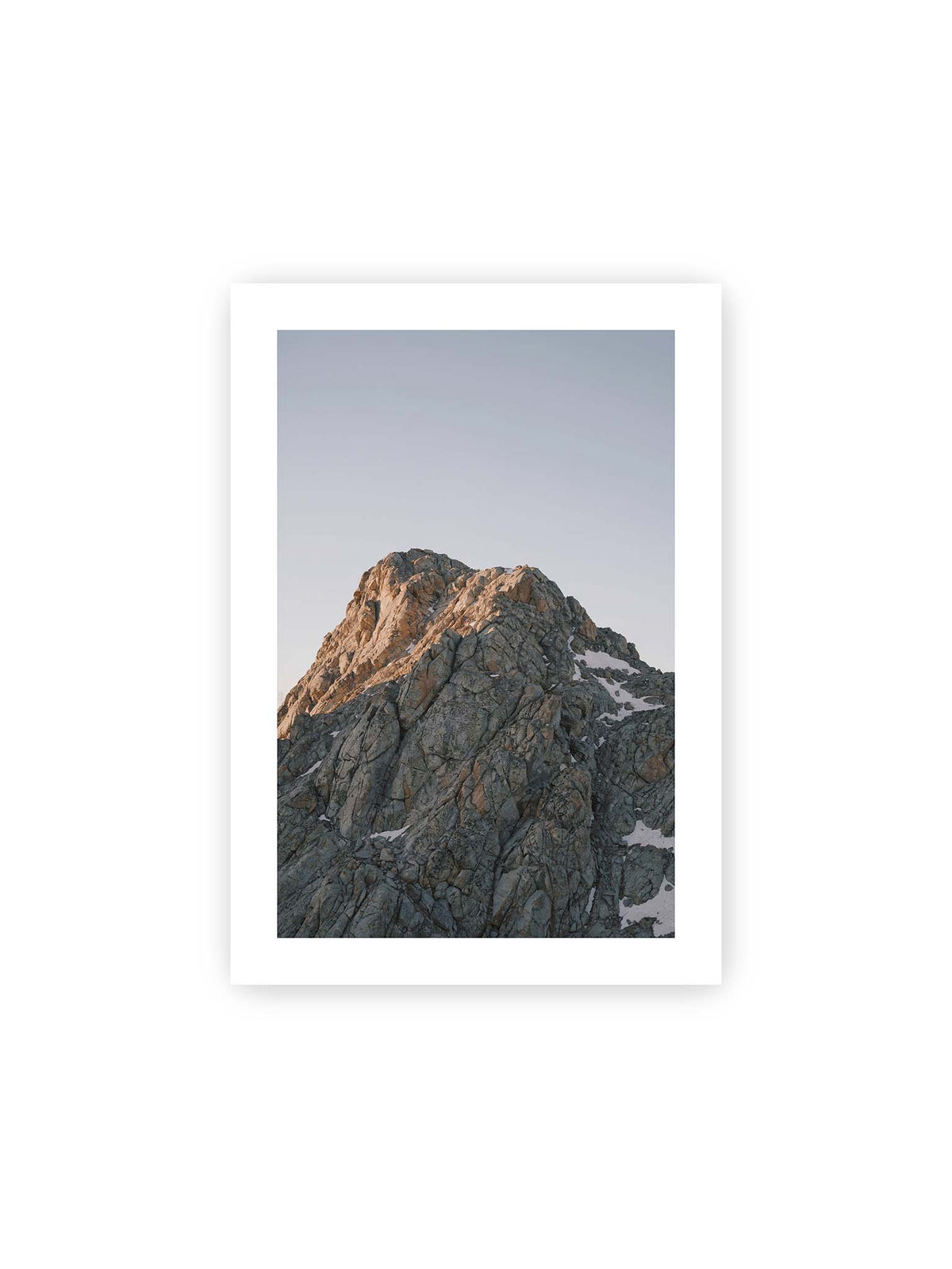 21x30 cm, weißer Galerie-Rand – Massiver grauer Felsgipfel mit kleinen Schneefeldern vor einem weiten blassblauen Himmel.
