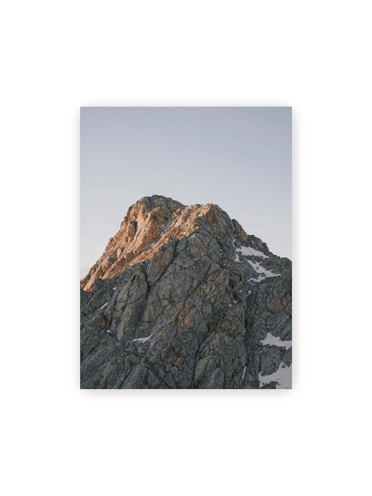 30x40 cm – Massiver grauer Felsgipfel mit kleinen Schneefeldern vor einem weiten blassblauen Himmel.