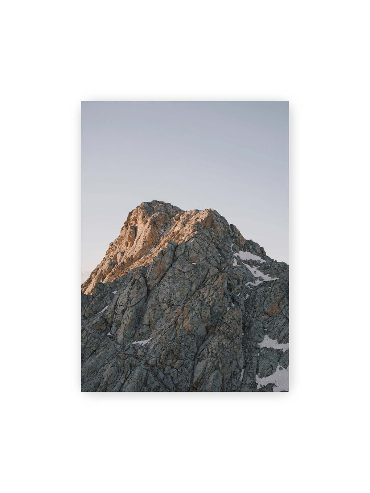 50x70 cm – Massiver grauer Felsgipfel mit kleinen Schneefeldern vor einem weiten blassblauen Himmel.
