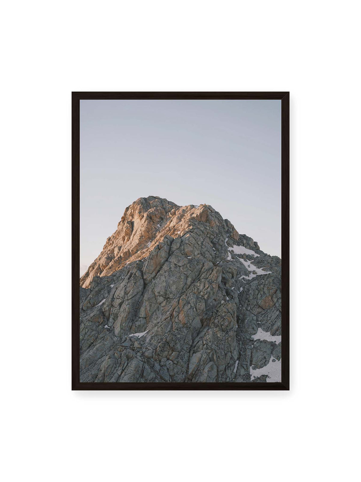 50x70 cm, schwarzer Rahmen – Massiver grauer Felsgipfel mit kleinen Schneefeldern vor einem weiten blassblauen Himmel.