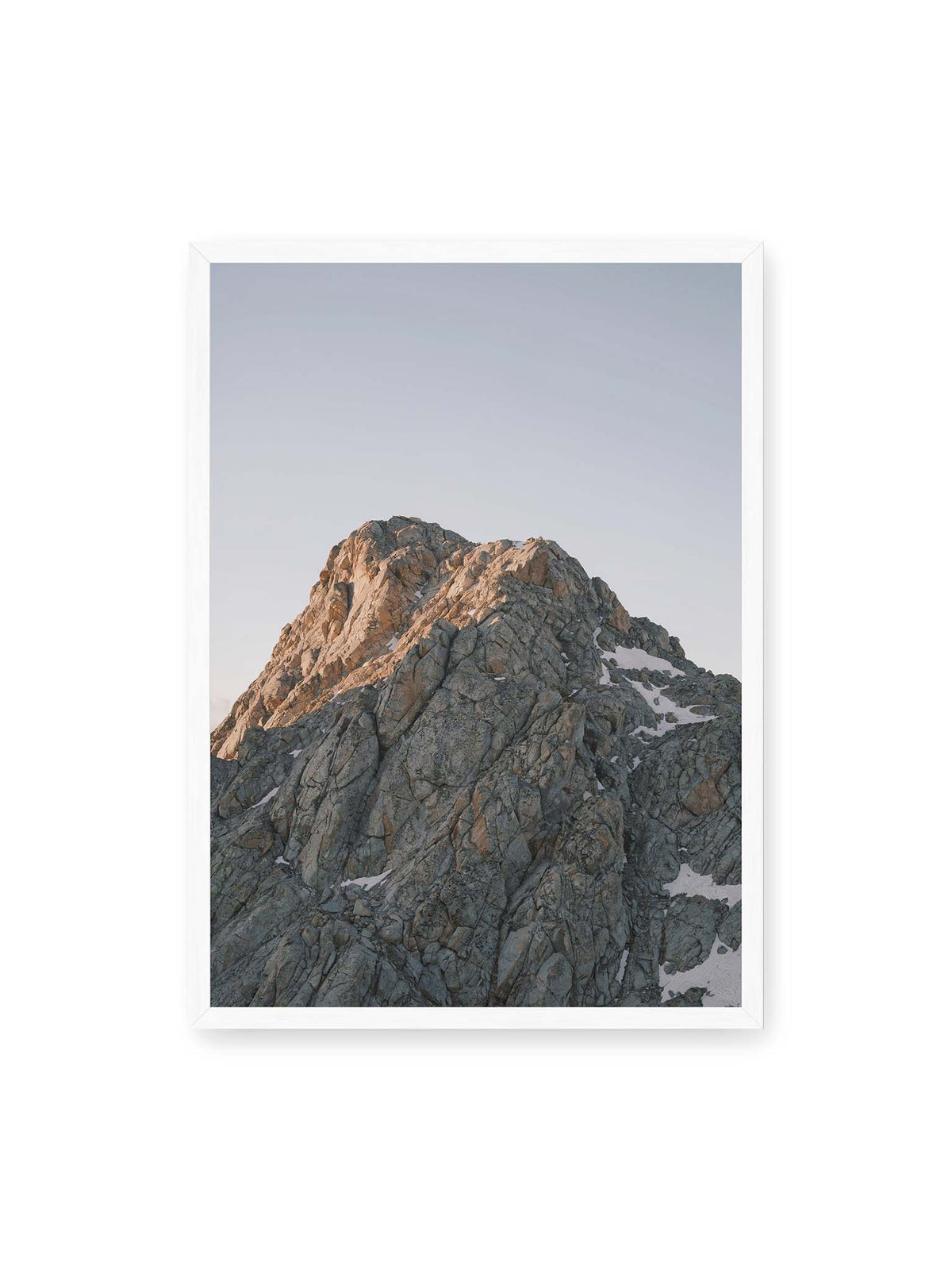 50x70 cm, weißer Rahmen – Massiver grauer Felsgipfel mit kleinen Schneefeldern vor einem weiten blassblauen Himmel.