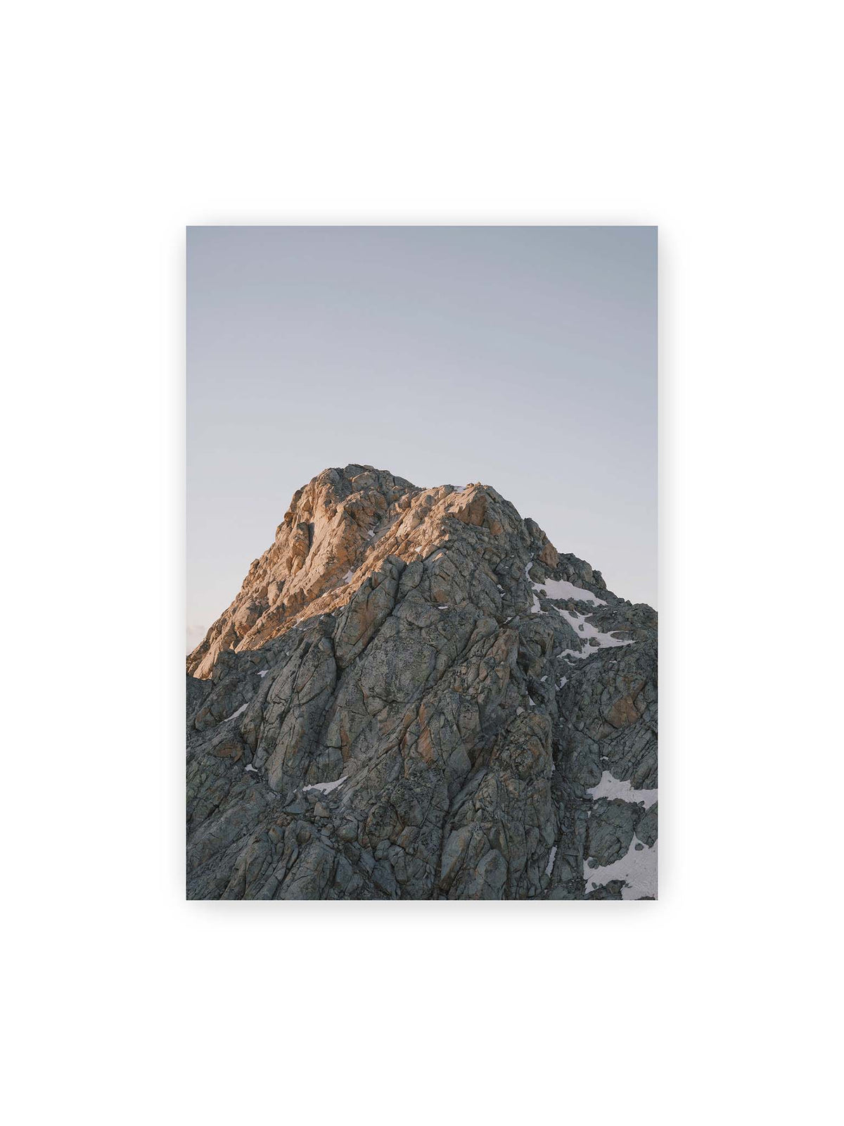 70x100 cm – Massiver grauer Felsgipfel mit kleinen Schneefeldern vor einem weiten blassblauen Himmel.
