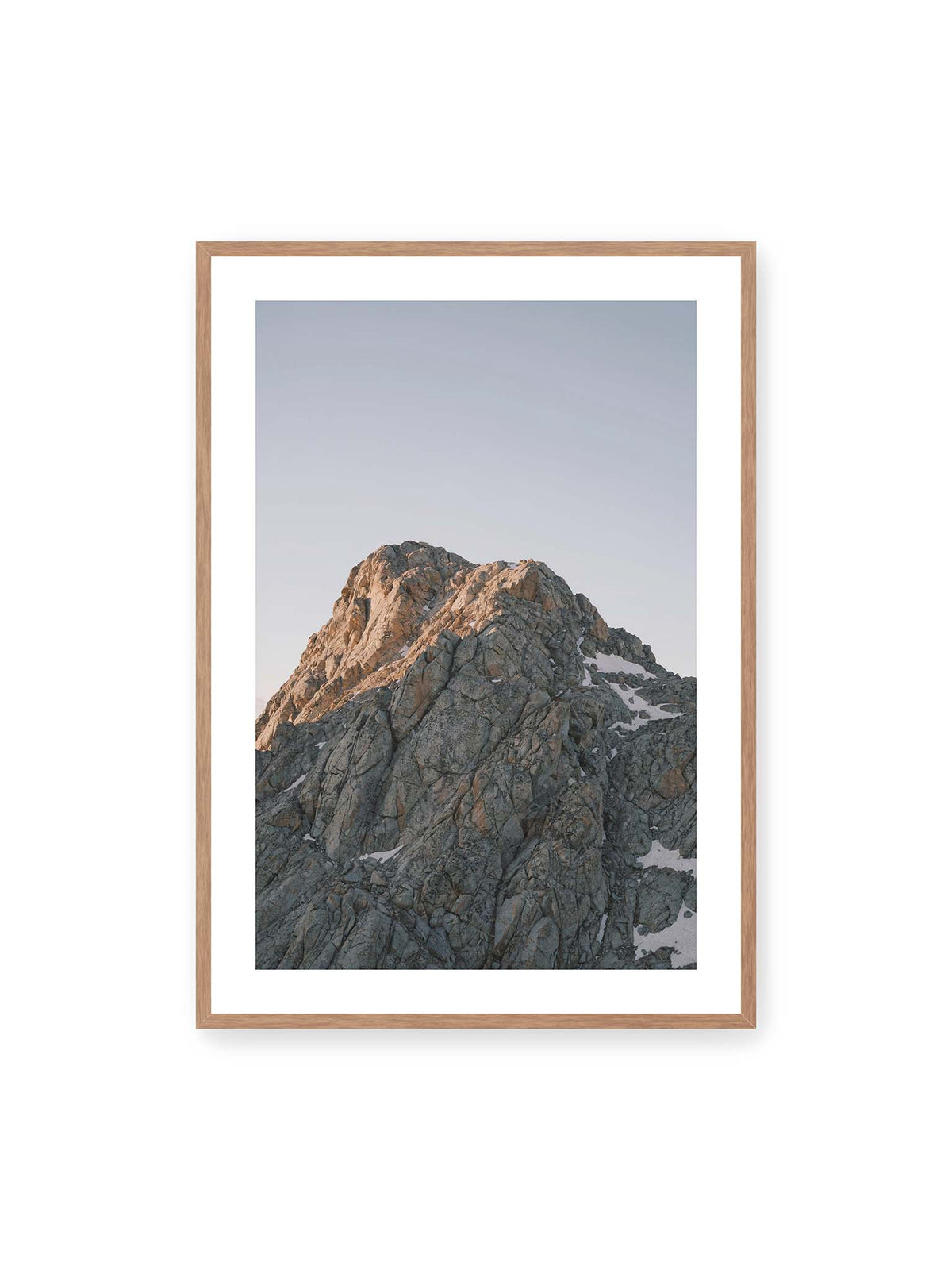 70x100 cm, weißer Galerie-Rand, Eichenrahmen – Massiver grauer Felsgipfel mit kleinen Schneefeldern vor einem weiten blassblauen Himmel.