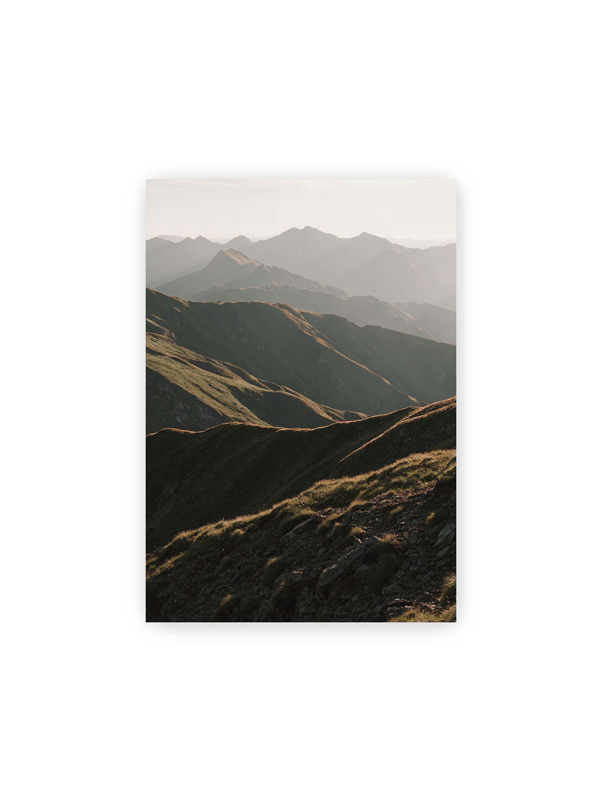 21x30 cm – Gestaffelte Bergketten mit grasbewachsenen Hängen und warm beleuchteten Konturen unter hellem Himmel.