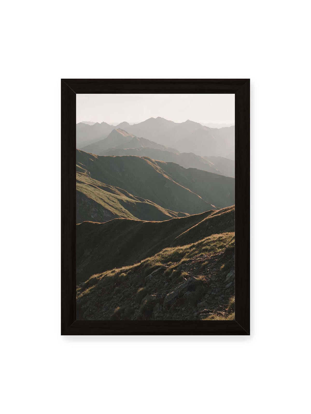 21x30 cm, schwarzer Rahmen – Gestaffelte Bergketten mit grasbewachsenen Hängen und warm beleuchteten Konturen unter hellem Himmel.